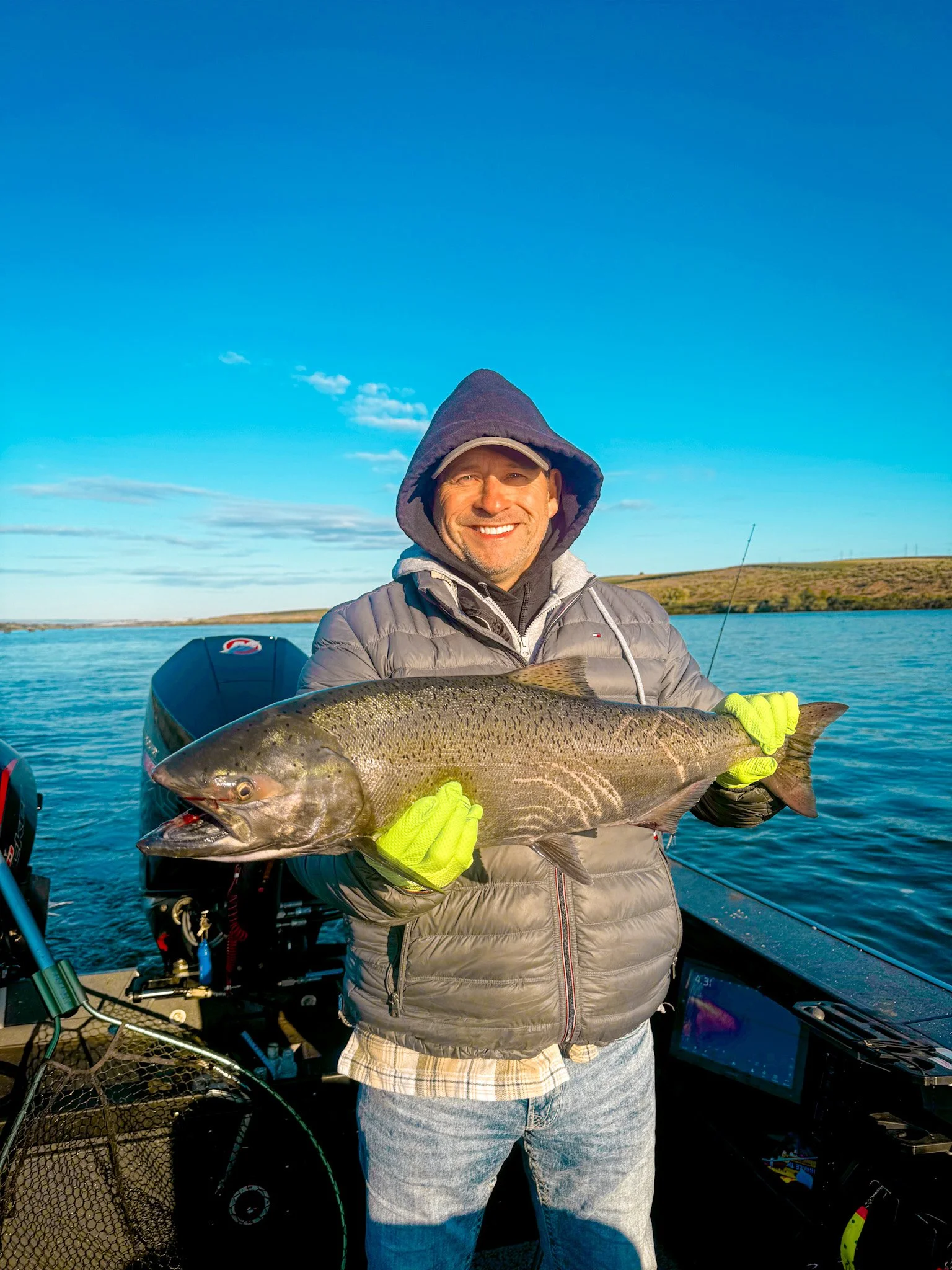 A man on a boat smiling while holding a large fish with a scenic lake and blue sky in the background.