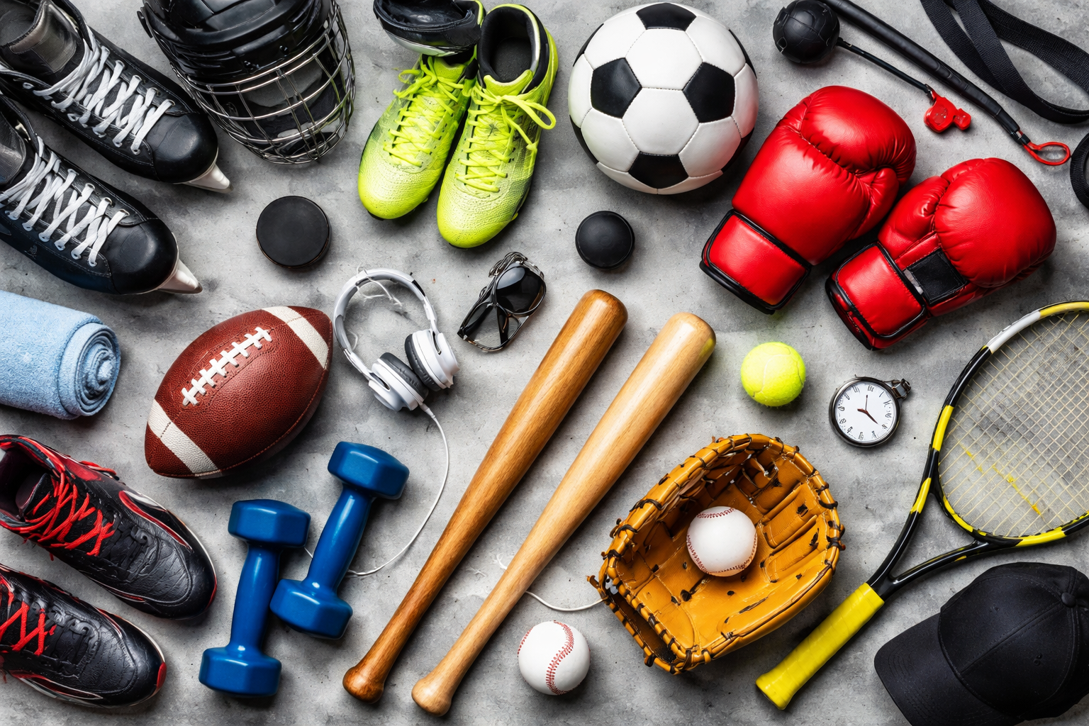 A collection of sports equipment including two pairs of ice skates, a pair of bright green soccer shoes, a soccer ball, red boxing gloves, a football, a tennis ball, a yellow tennis racket, a baseball glove with a baseball, blue dumbbells, a pair of wooden baseball bats, a baseball, a pair of headphones, a stopwatch, a black cap, a rolled blue towel, black hockey gloves, a lacrosse stick, a pair of sunglasses, and a black water bottle on a concrete surface.