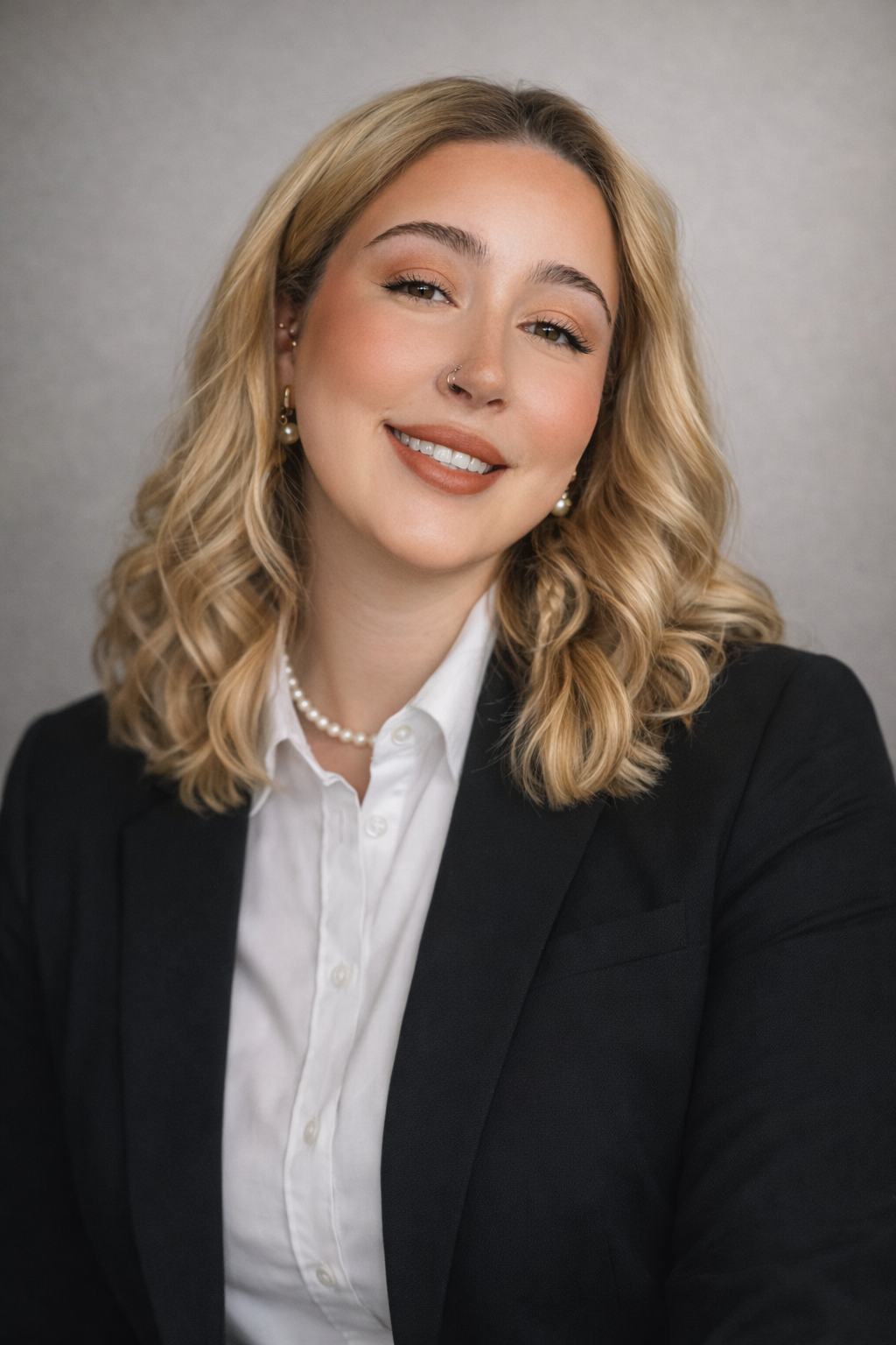 A professional woman with blonde wavy hair, wearing a black blazer, white shirt, pearl necklace, and pearl earrings, smiling in front of a plain gray background.