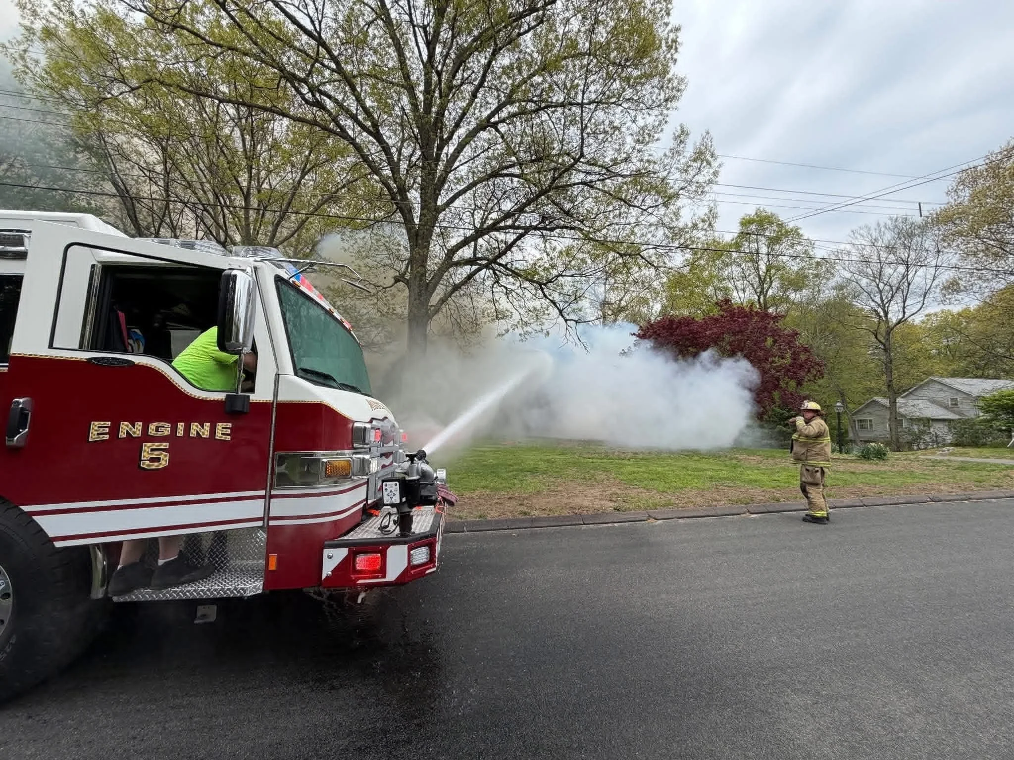 Firefighters using a hose to spray water on a fire, with smoke rising from a tree in a neighborhood.