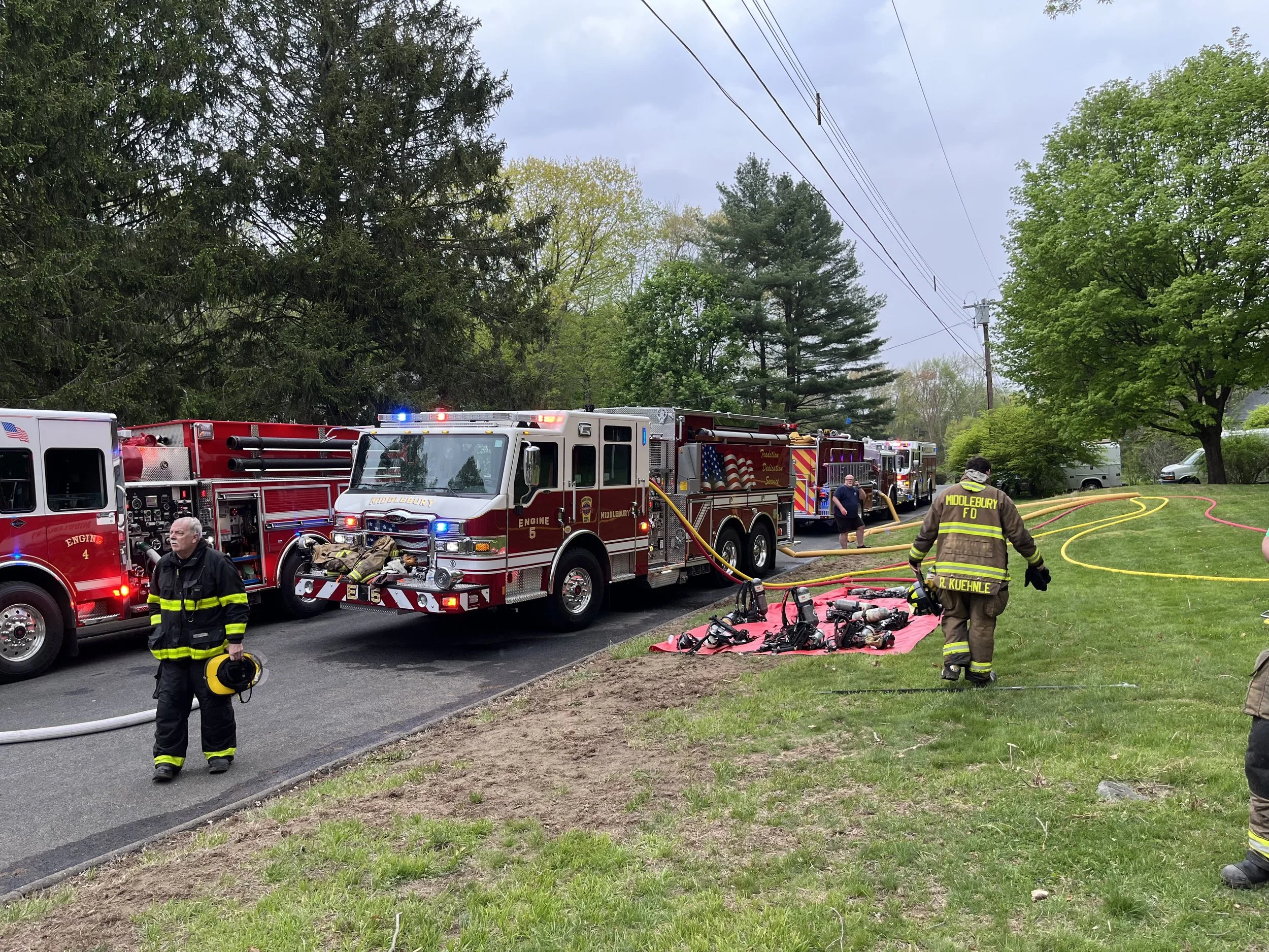 Firefighters and fire trucks at the scene of an emergency on a residential street, with firefighting equipment and hoses laid out on the grass.