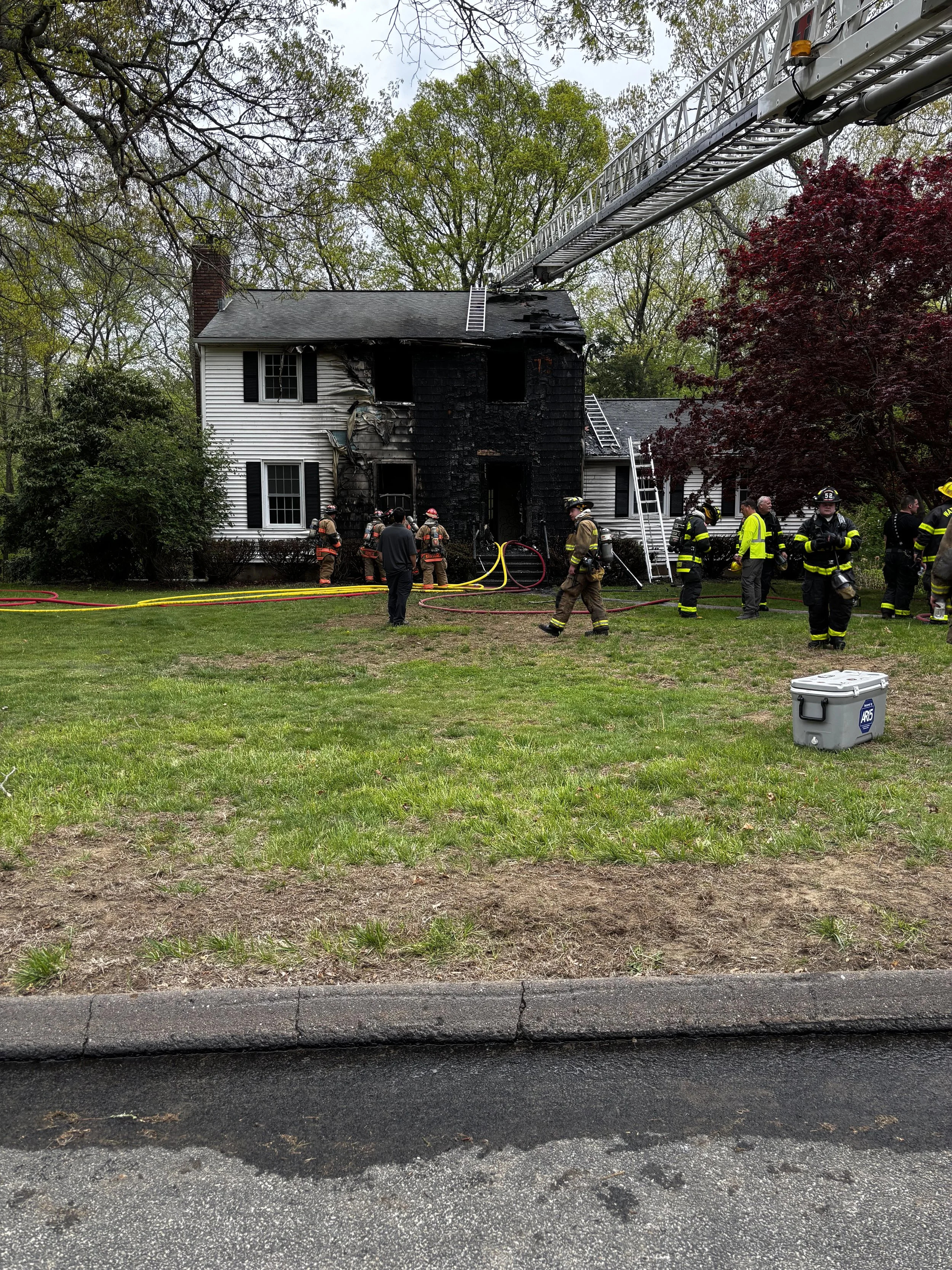 Fire-damaged house with firefighters and equipment outside, including ladders and a cooler.