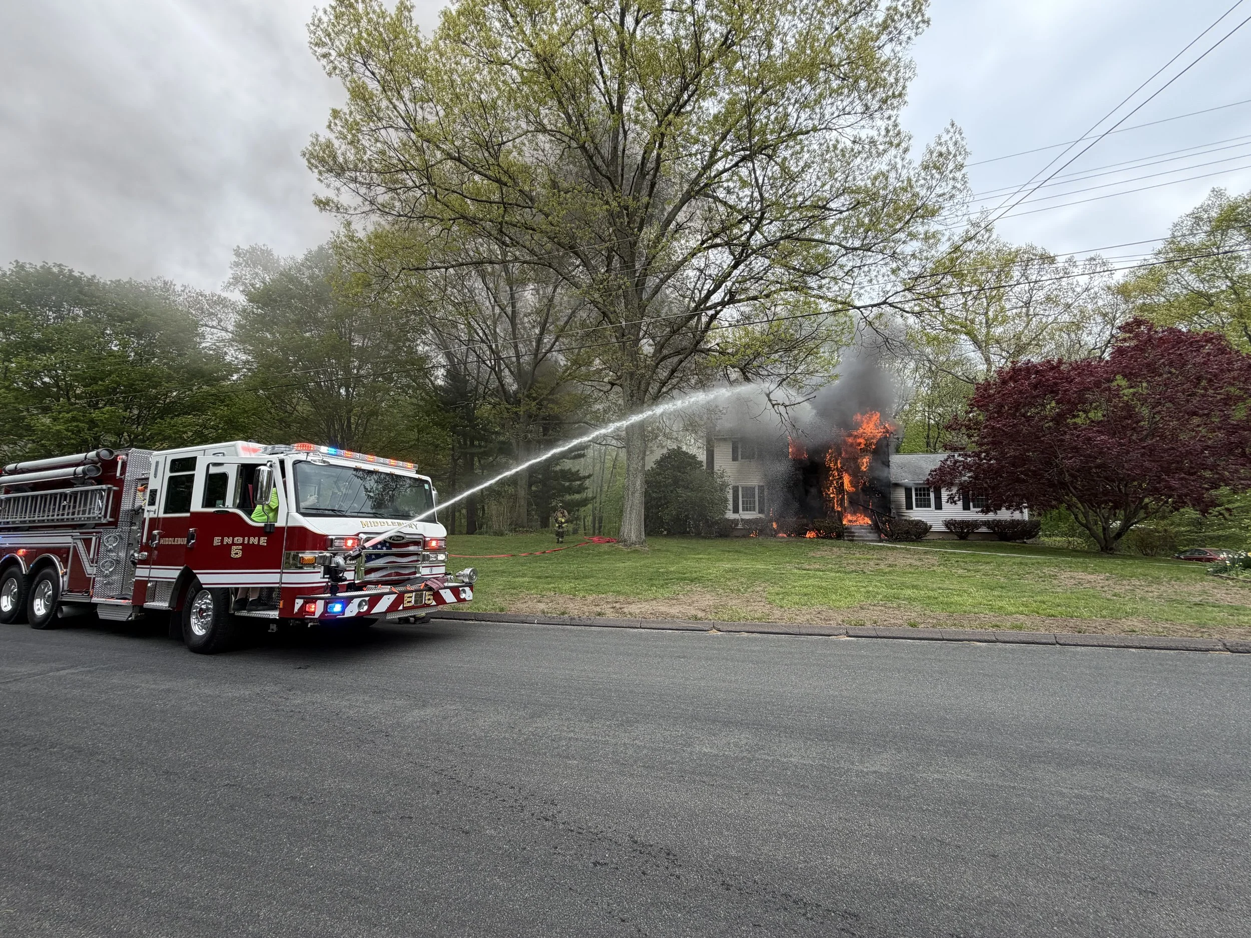 Firefighters spraying water on a house fire with flames and smoke coming out of the house, with a fire truck in front and trees around.