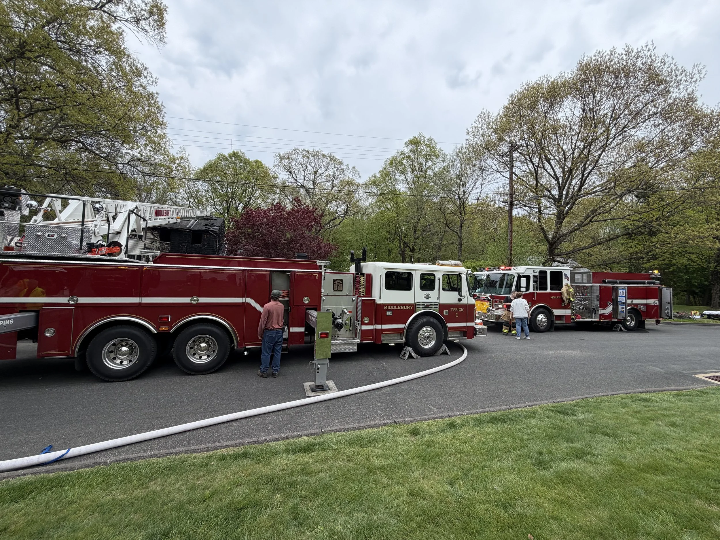 Fire trucks and firefighters parked on the street with trees and cloudy sky overhead.