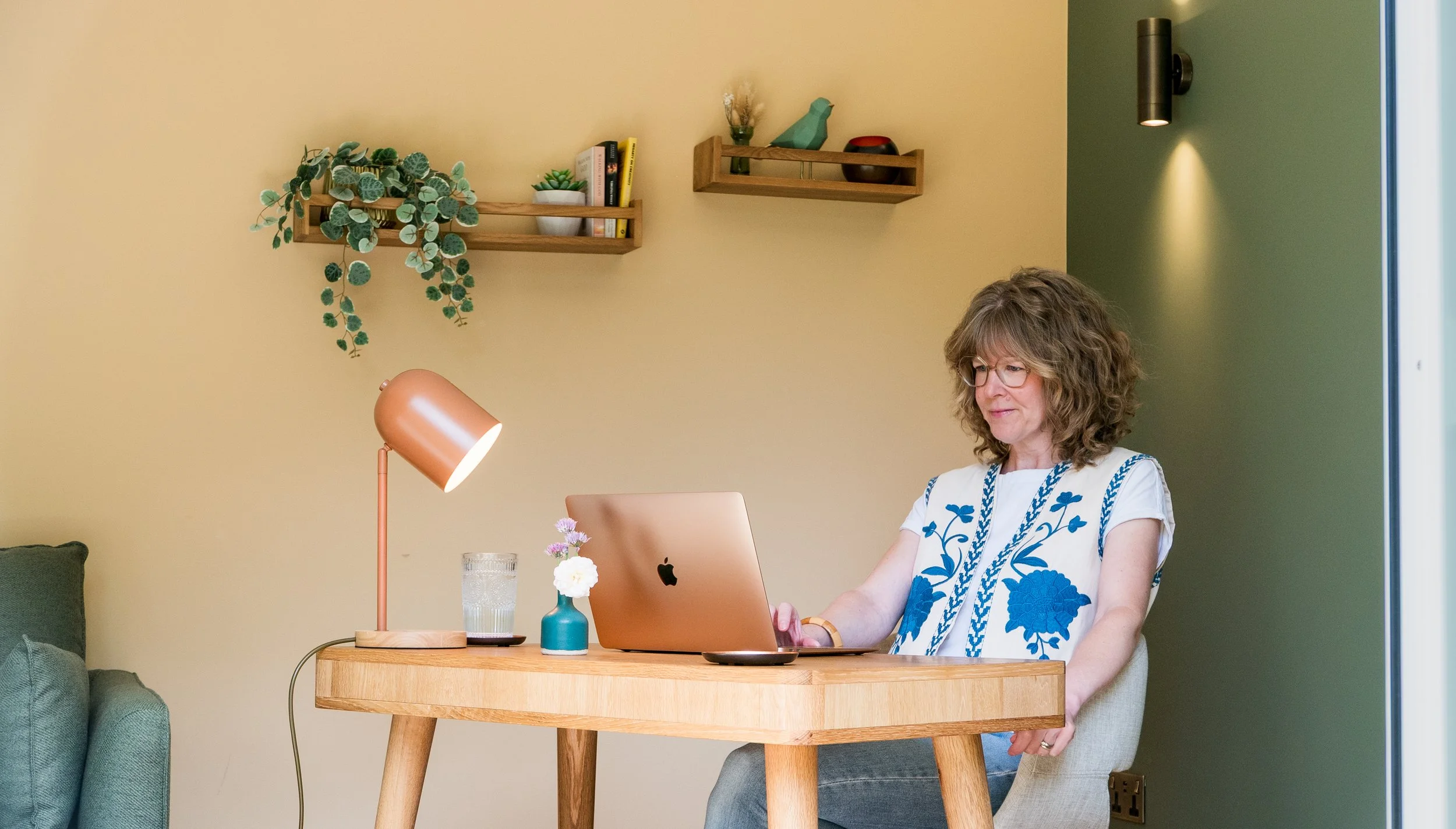 Woman sitting at a wooden table working on a laptop, with a glass of water, small flower vase, and pink desk lamp. Shelves with plants and decor on beige and green walls.