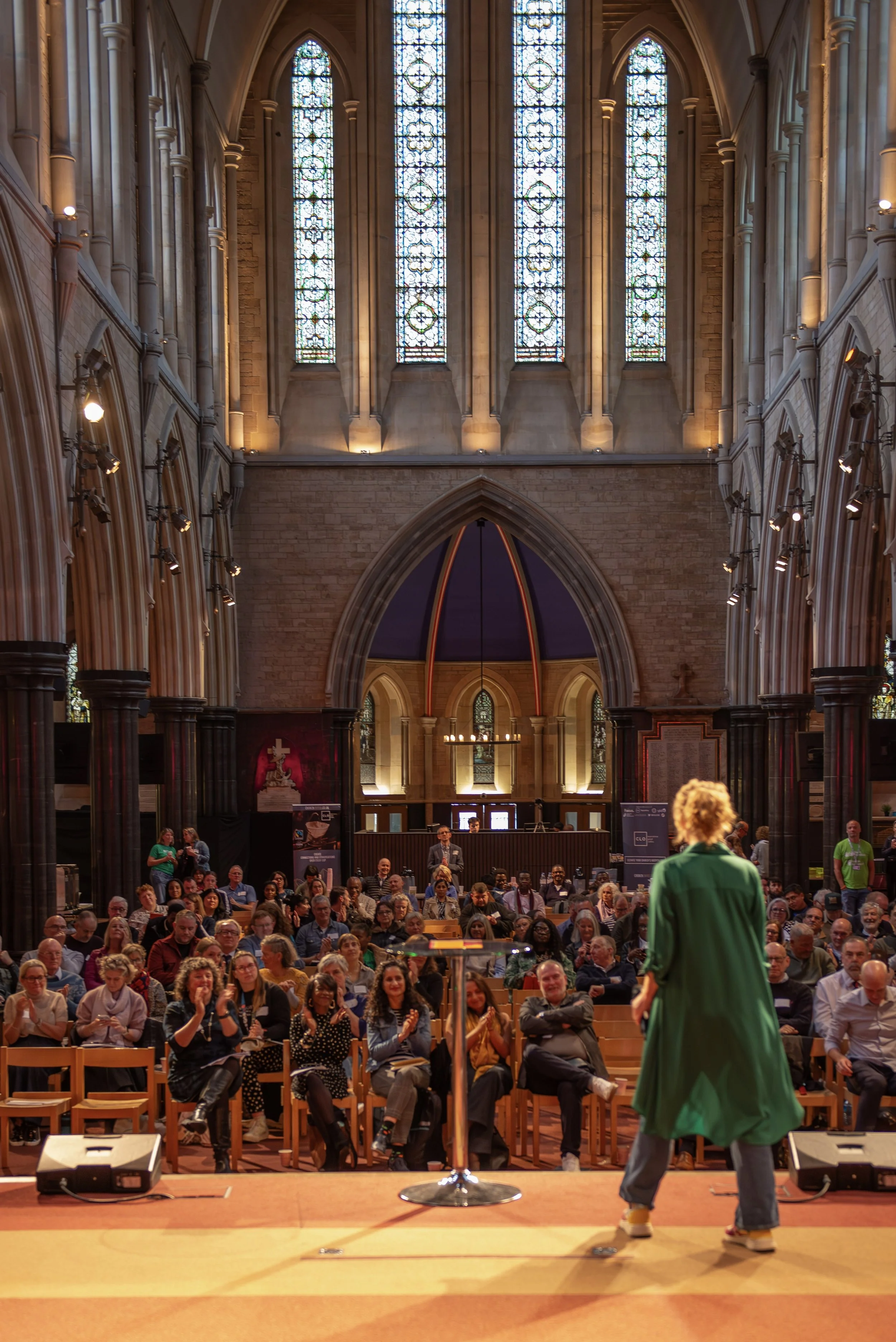 A woman in a green coat speaking on stage in front of an audience inside a large historic church or cathedral with stained glass windows.