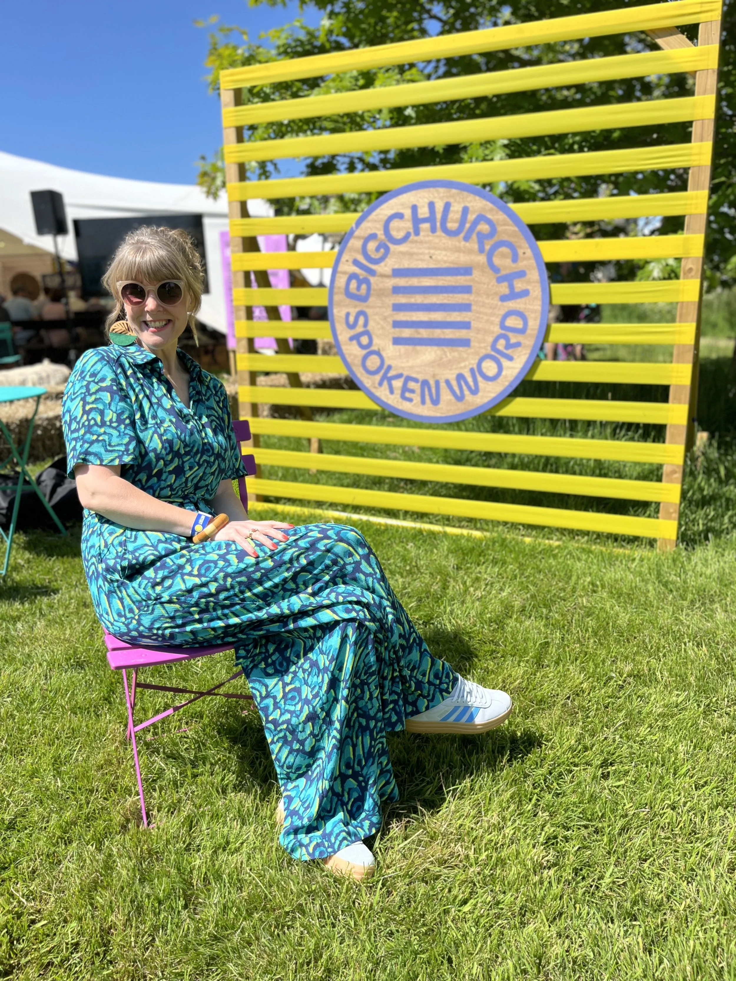 A woman wearing sunglasses and a blue patterned dress sitting on a pink chair outdoors near a yellow fence and a sign that reads 'SPOKES BIG CHURCH POKEN WORD CH.' She is smiling at the camera on a sunny day with a tent and people in the background.