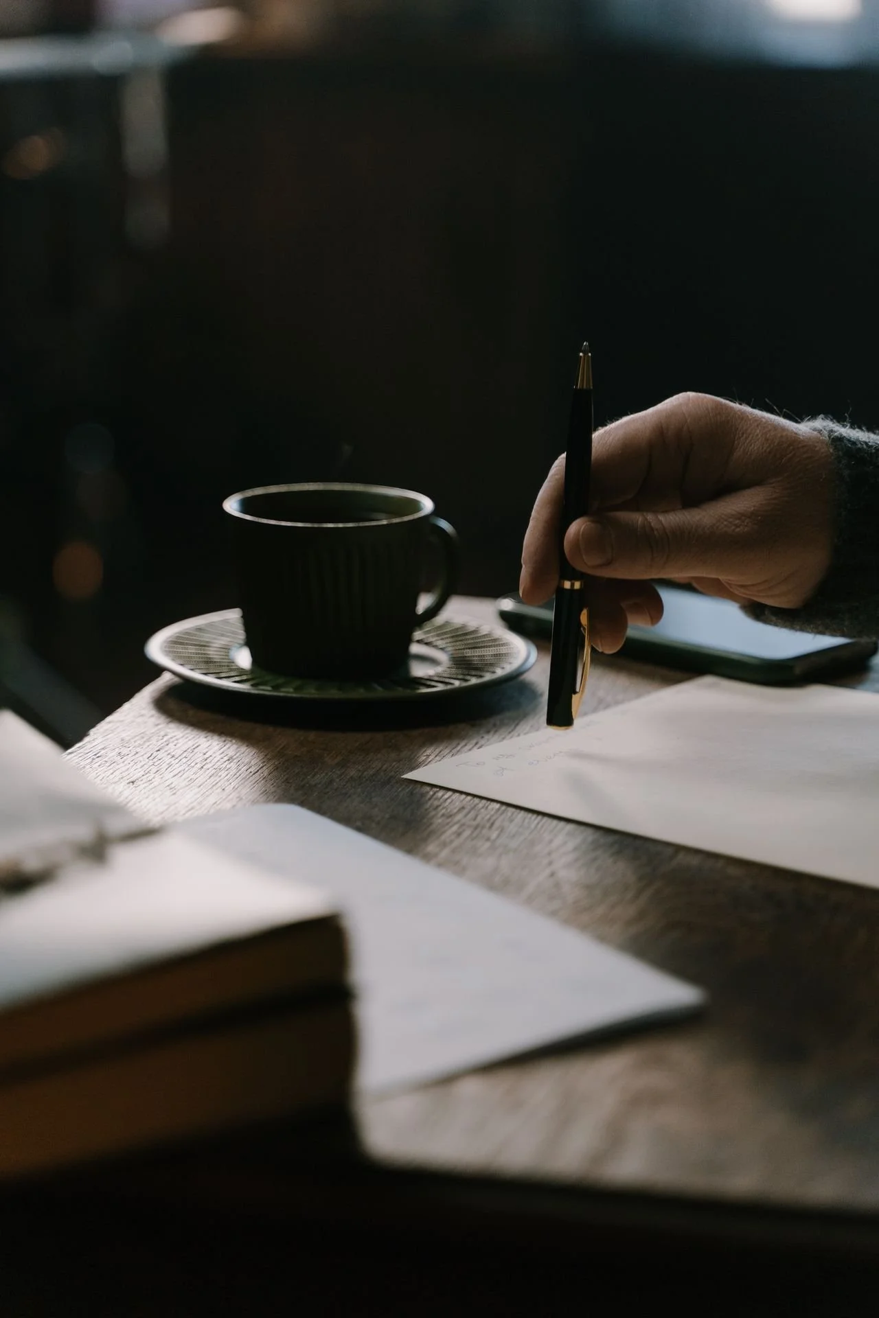 Hand holding a pen and writing on paper on a wooden table, with a black coffee mug on a saucer nearby and books in the foreground.