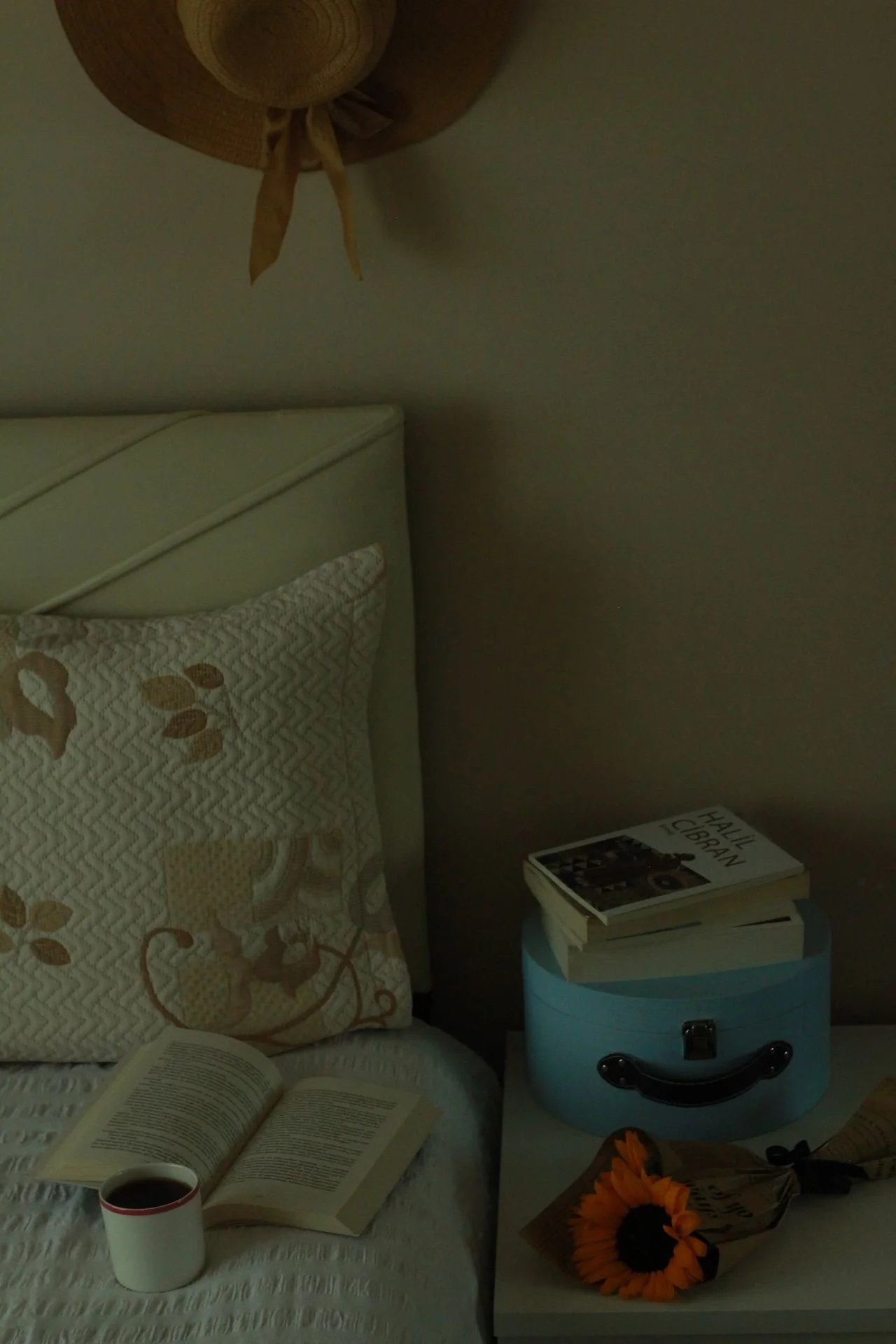 A cozy bedroom corner with a bed, quilted pillow with floral design, an open book, a cup of coffee, a small white table with a sunflower, a blue decorative box, some books including 'Halle Cibrian', umbrella, and a pair of shoes, against a beige wall with a ceiling fan.