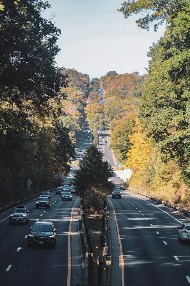 A highway with cars traveling in both directions surrounded by trees with fall foliage, extending up a rolling hill.