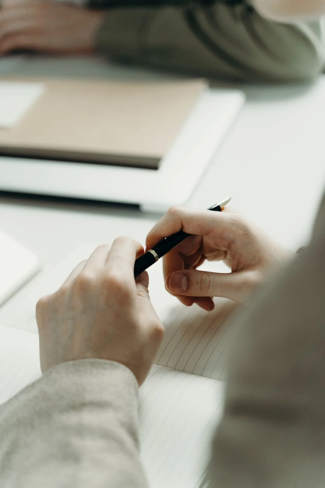 Close-up of a person's hands holding a black pen and writing in a notebook with lined pages.