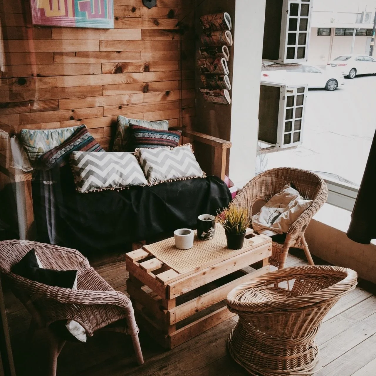 Cozy indoor seating area with wooden and wicker furniture, patterned pillows, a wooden pallet coffee table with potted plants and cups, and window with open shutters looking outside at parked cars.