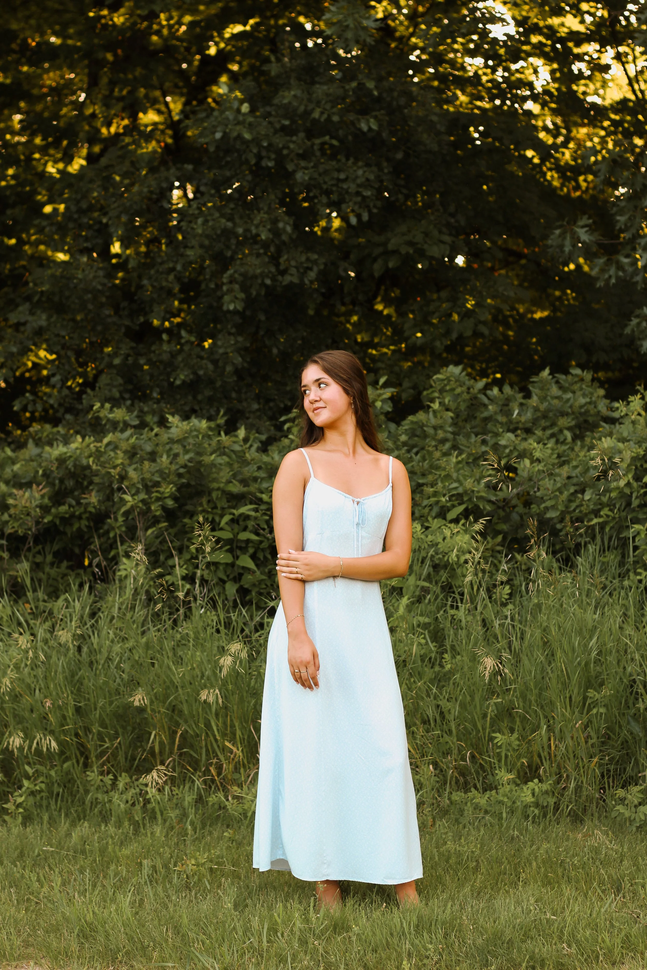 A young woman in a light blue dress standing in a grassy field with green bushes and tall trees in the background, illuminated by warm sunlight.