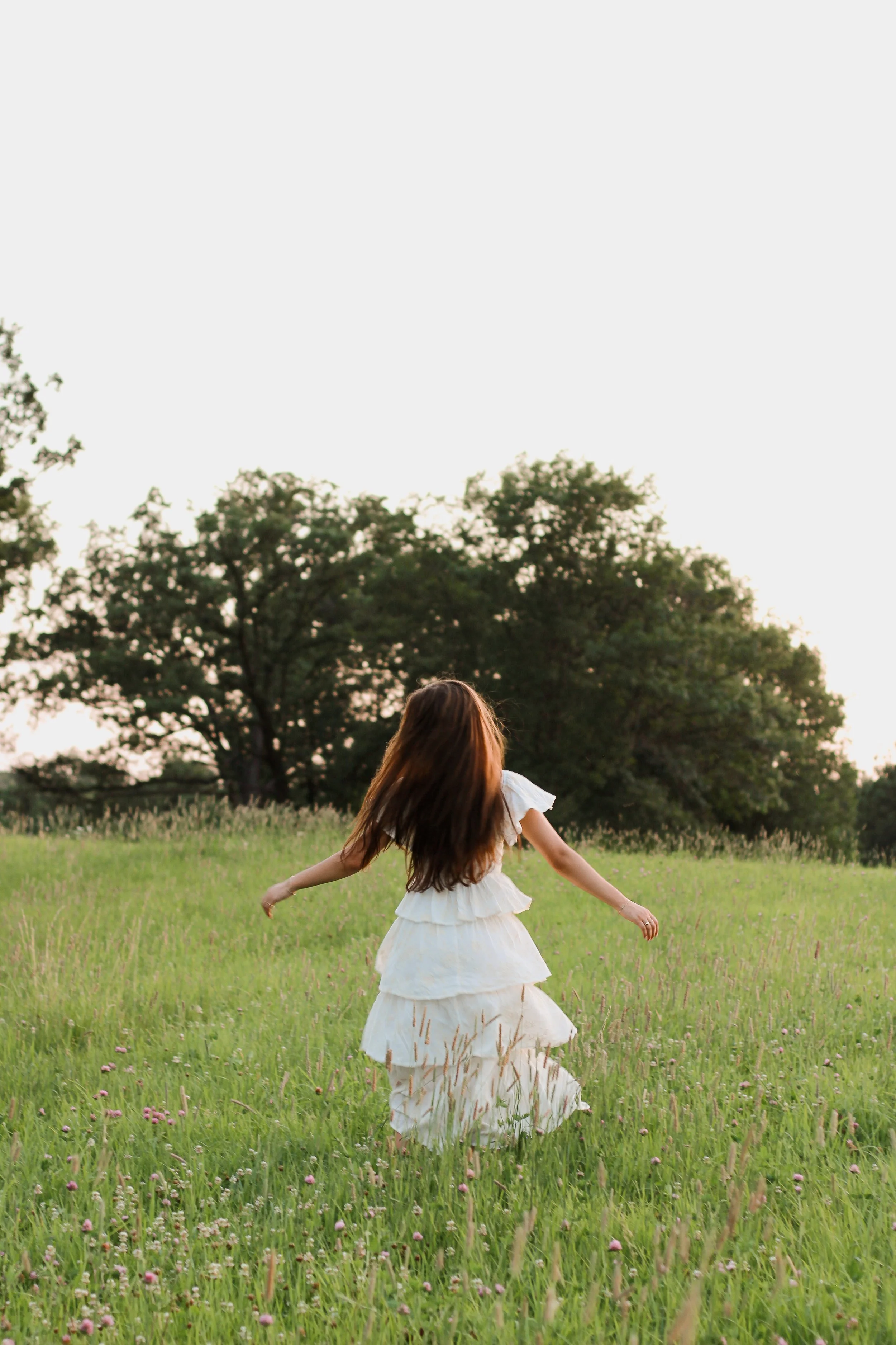A woman in a white dress spinning in a green field with trees in the background at sunset.
