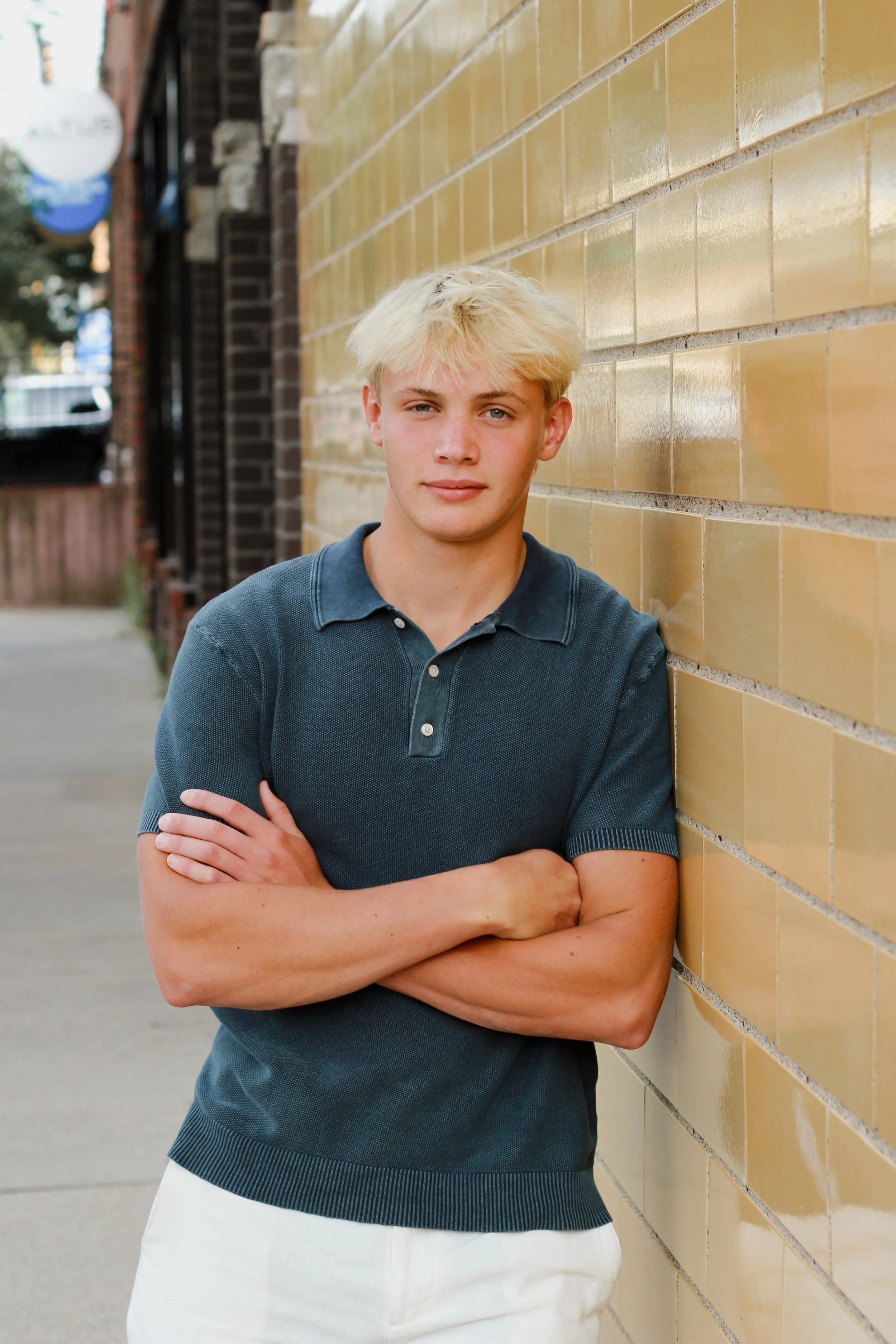 A young man with blond hair and blue eyes leaning against a yellow brick wall outside, crossing his arms and looking at the camera.
