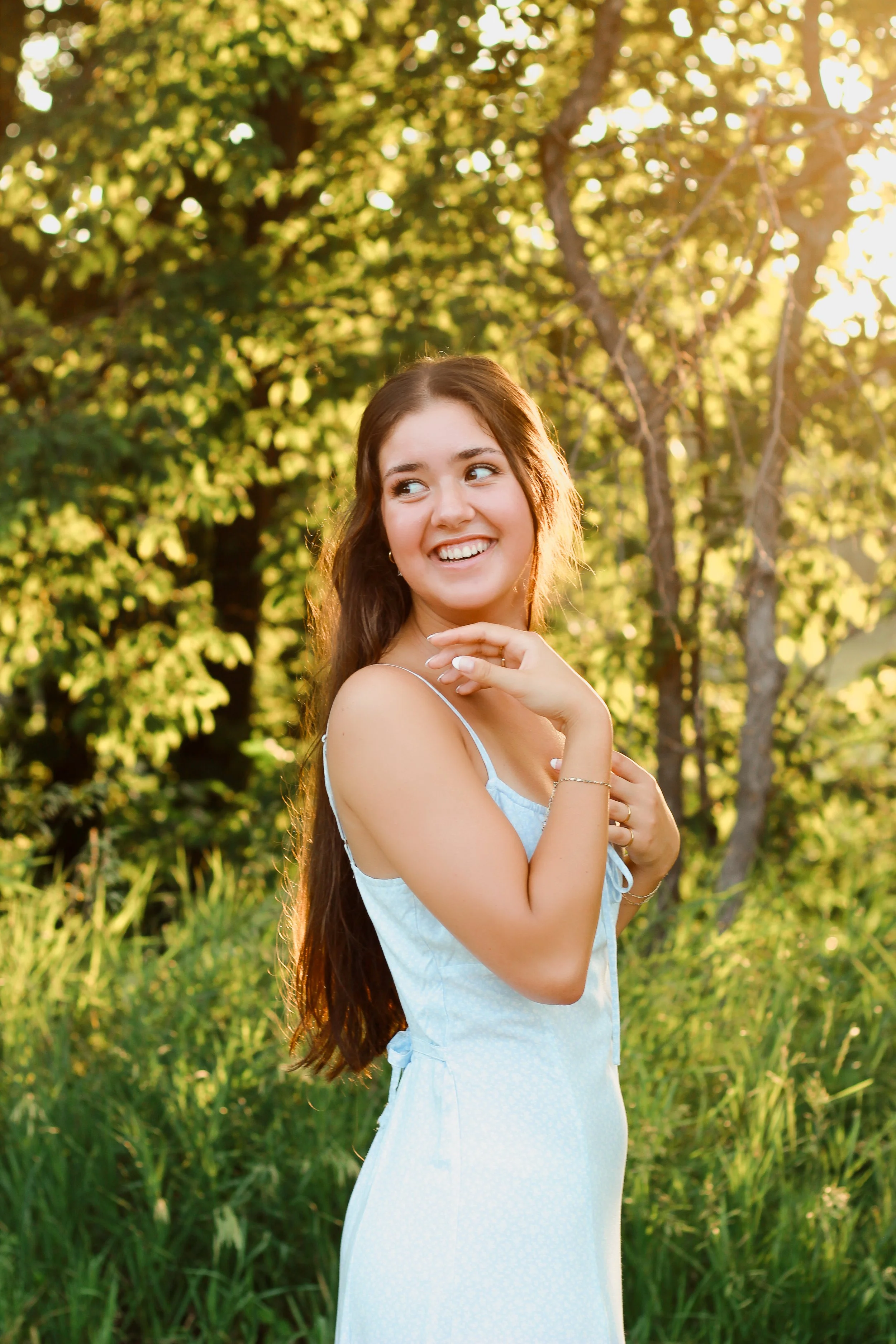 A young woman with long brunette hair wearing a light blue dress, smiling and looking to her right, standing outdoors with green trees and grass in the background, illuminated by warm sunlight.