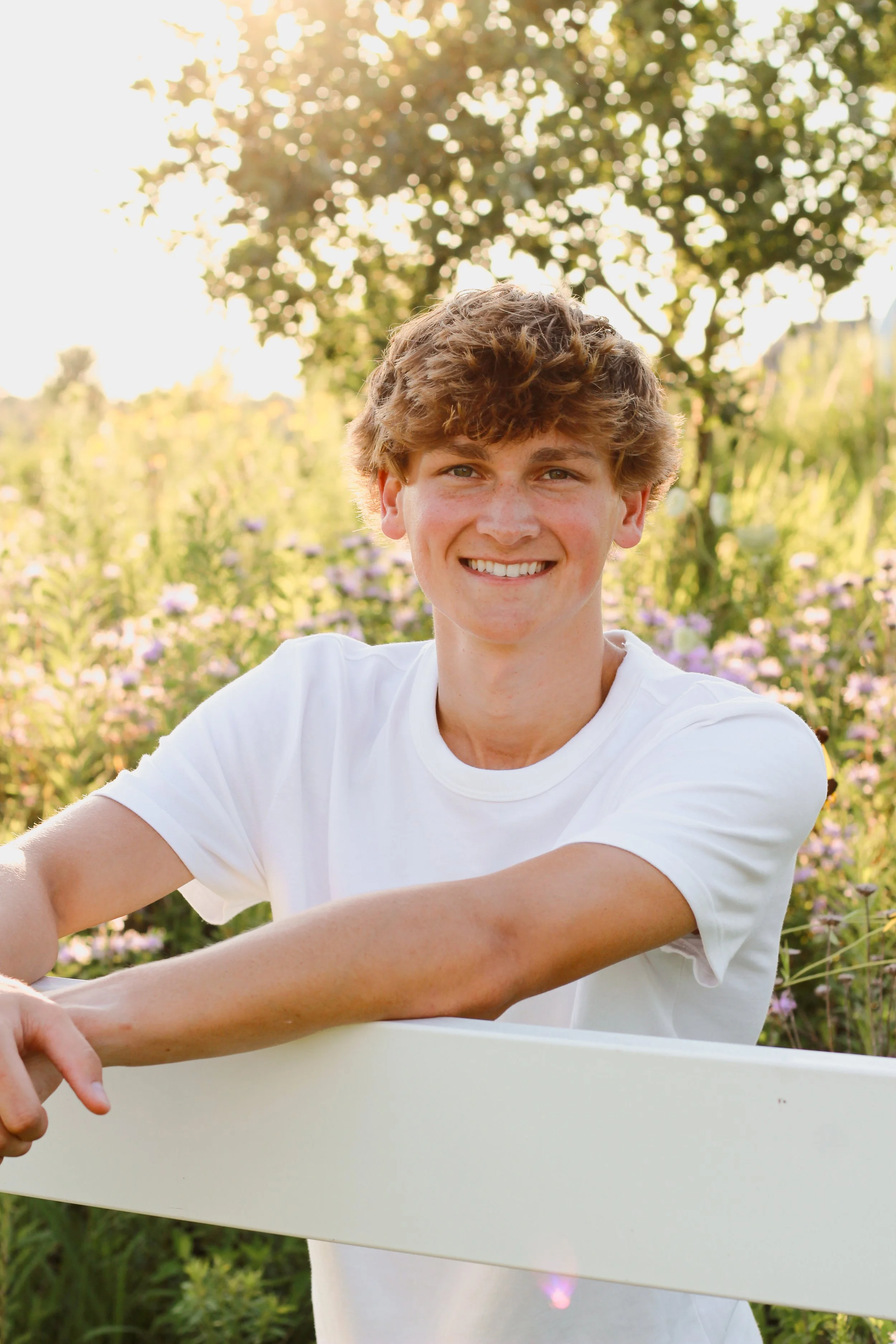 A smiling young man with curly hair in a white T-shirt leans on a white railing outdoors in a garden with purple flowers and green trees, illuminated by warm sunlight.