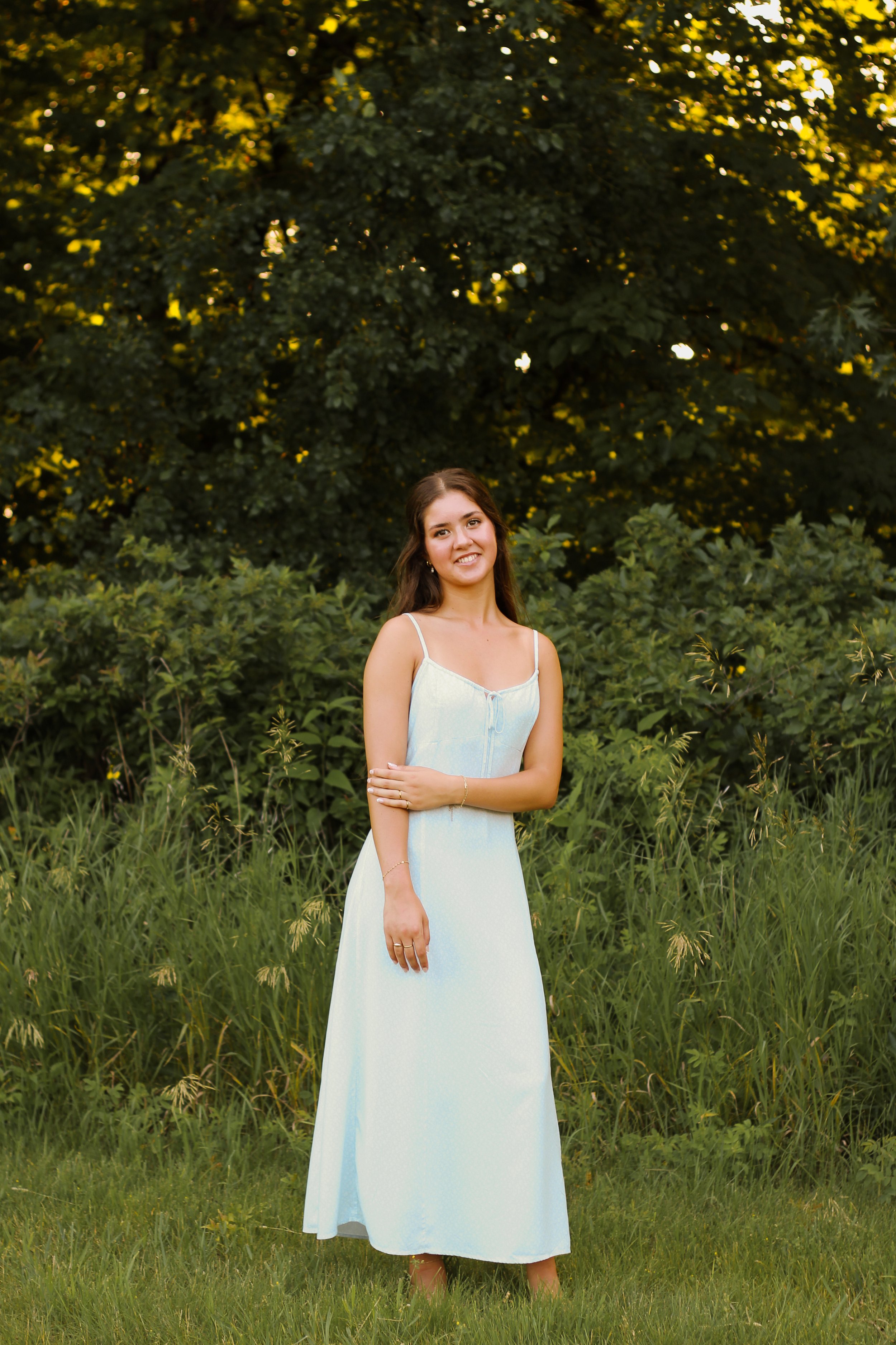 A young woman in a white dress standing outdoors on grass with a backdrop of dense green bushes and trees at sunset.