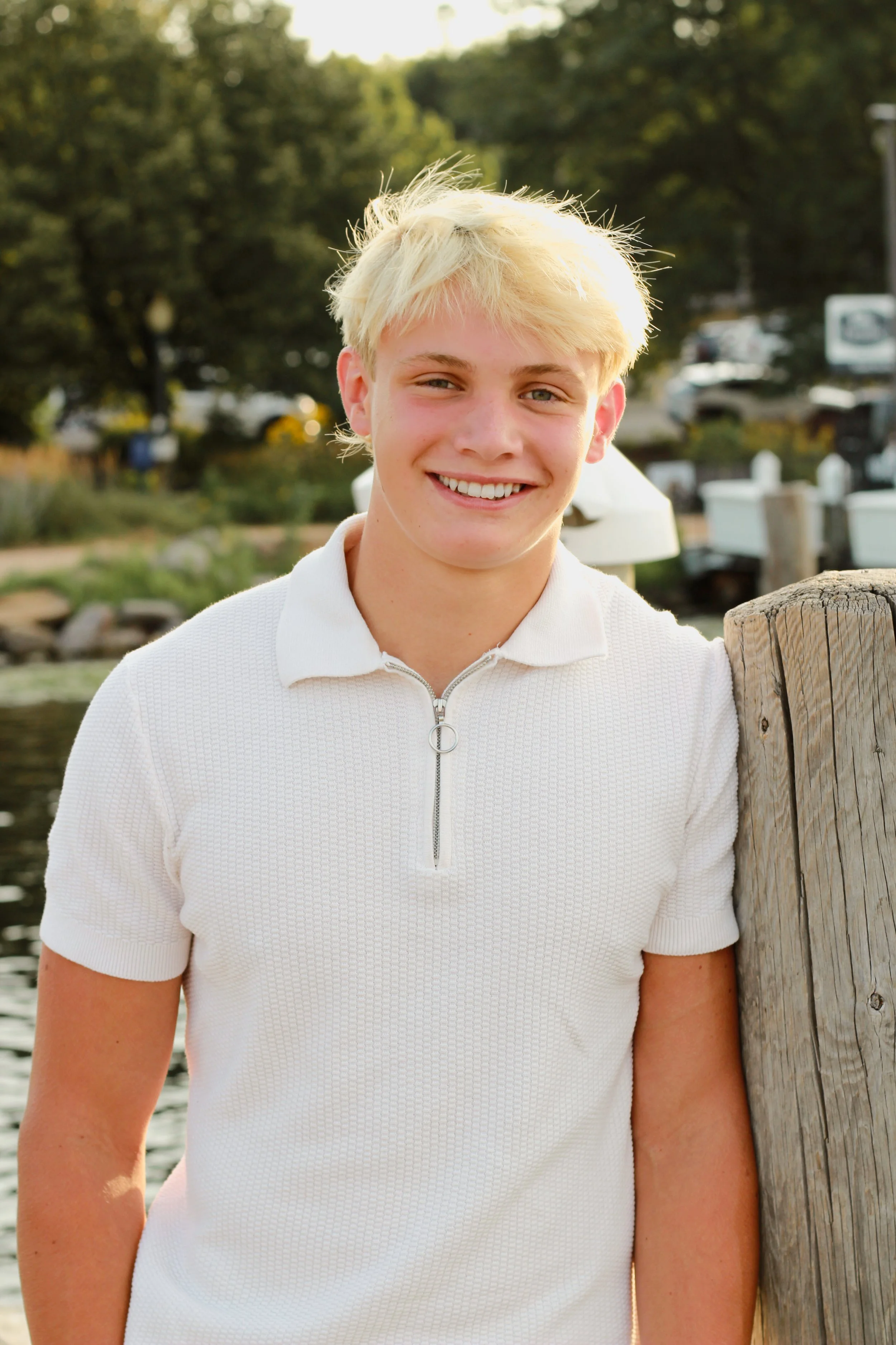 A young man with blonde hair smiling outdoors near a wooden post, with trees and parked cars in the background during the daytime.