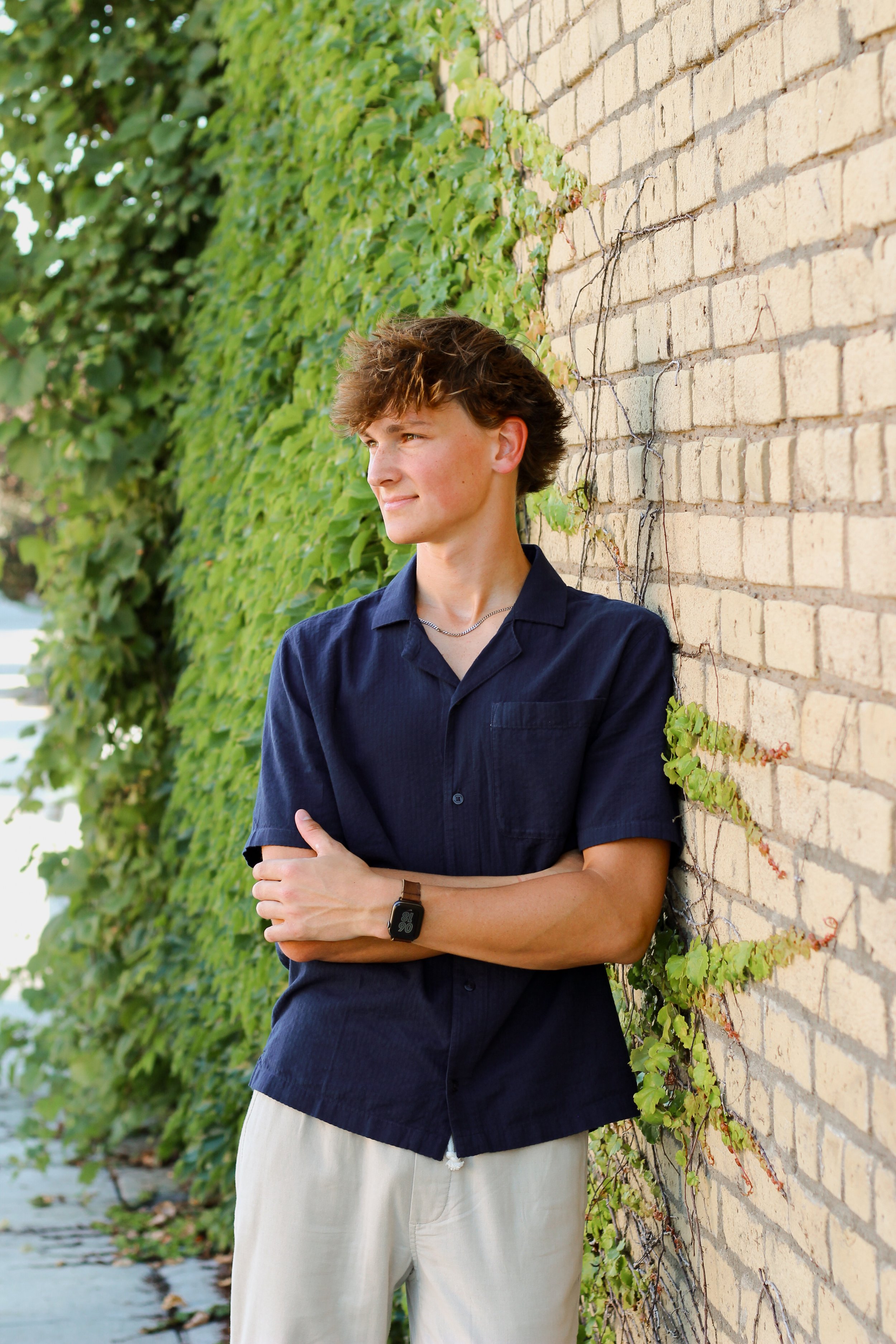 Young man with short, curly brown hair, wearing a navy blue short-sleeved button-up shirt, light-colored pants, a smartwatch, and a silver chain necklace, leaning against a brick wall with green ivy plants.