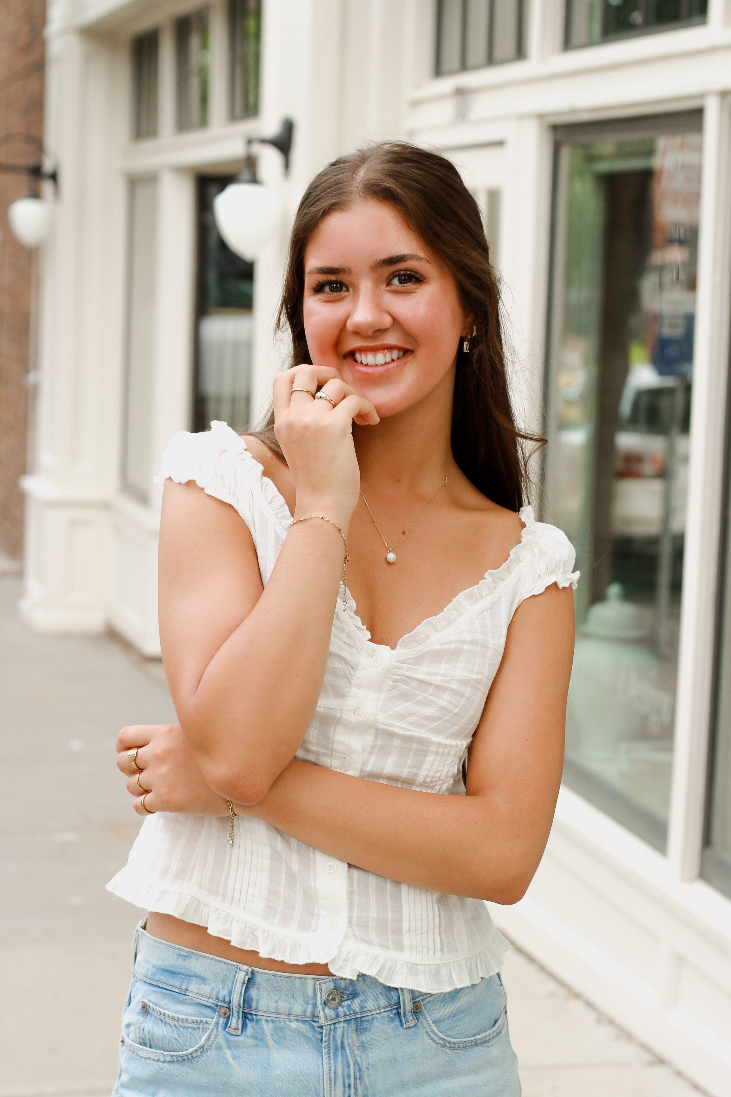 Young woman with long brown hair smiling outdoors, wearing a white sleeveless top and denim jeans, standing in front of a building with large windows.