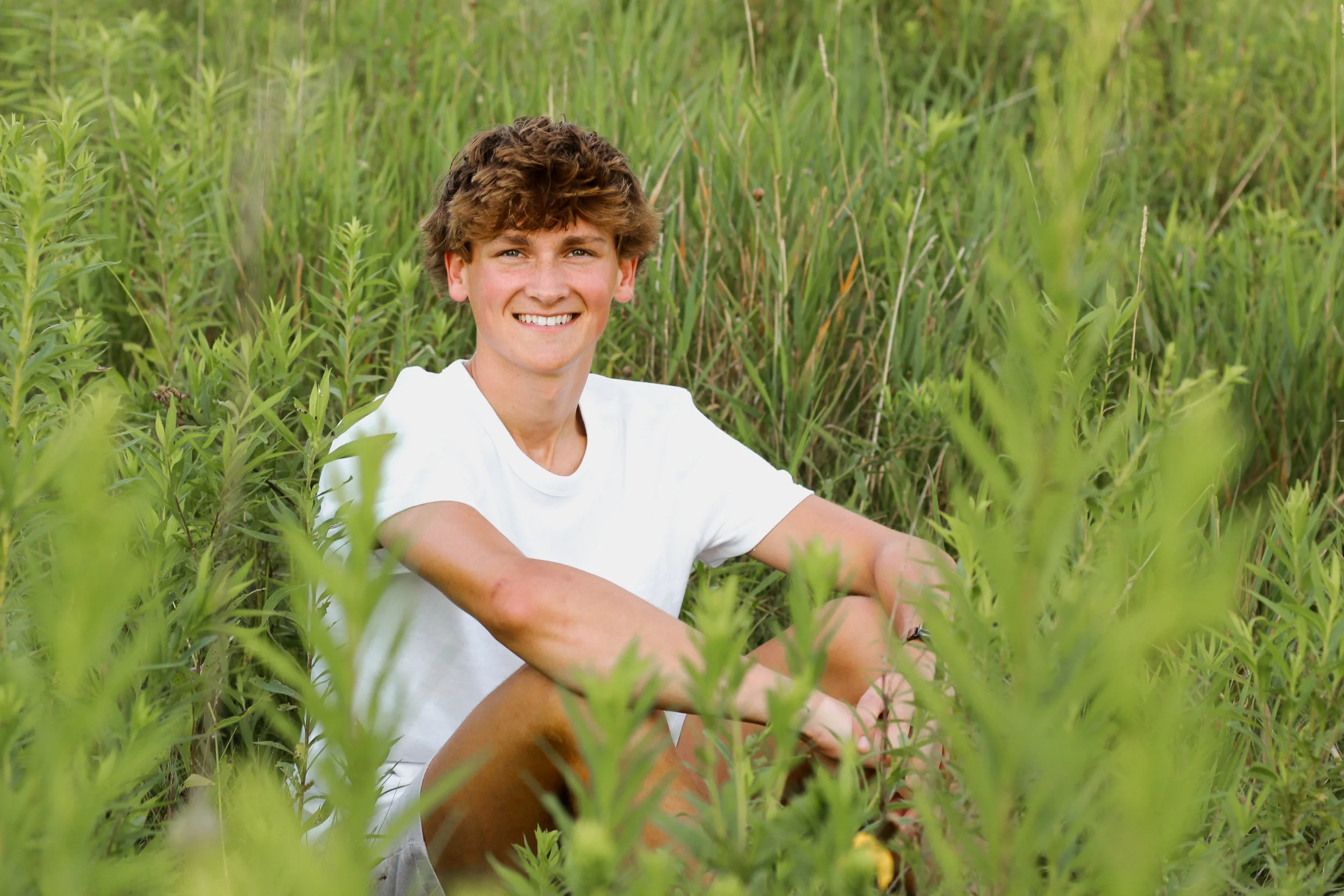 Young man sitting in a lush green field of tall plants, smiling at the camera.