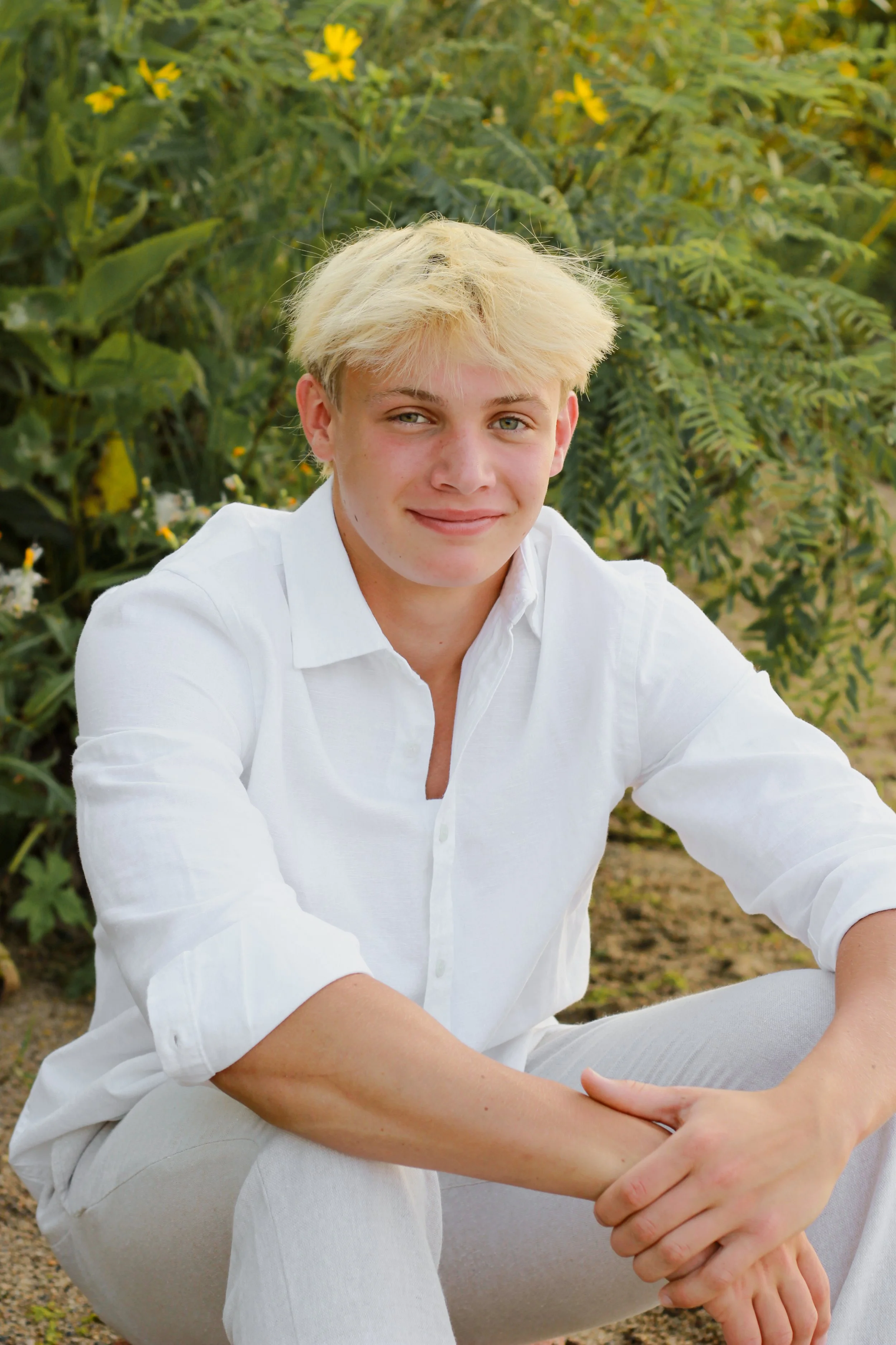 A young person with blonde hair wearing a white shirt sitting outdoors in front of green plants and yellow flowers.