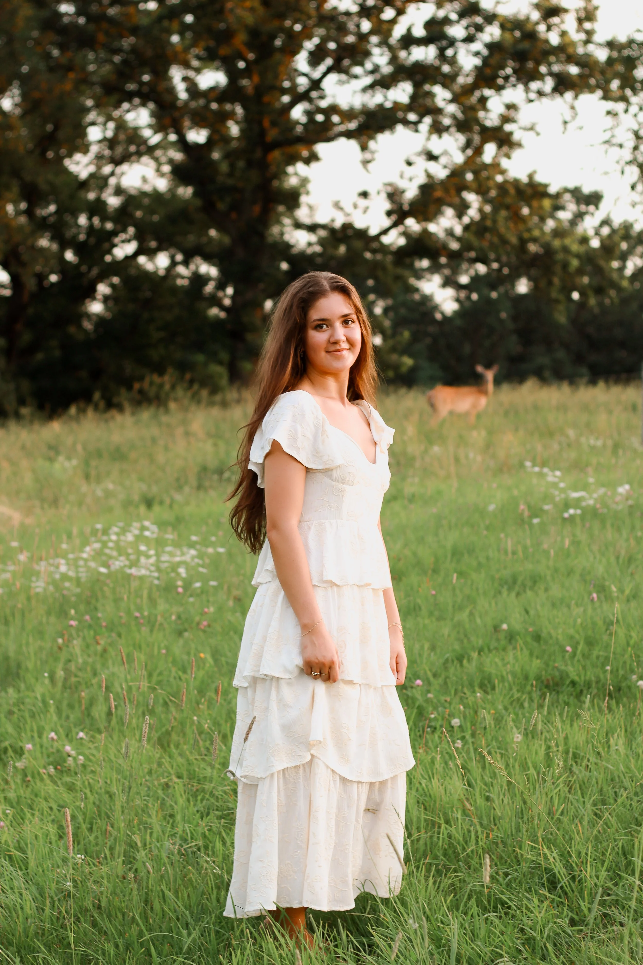 Young woman in a cream-colored dress standing in a green field with trees in the background and deer in the distance.