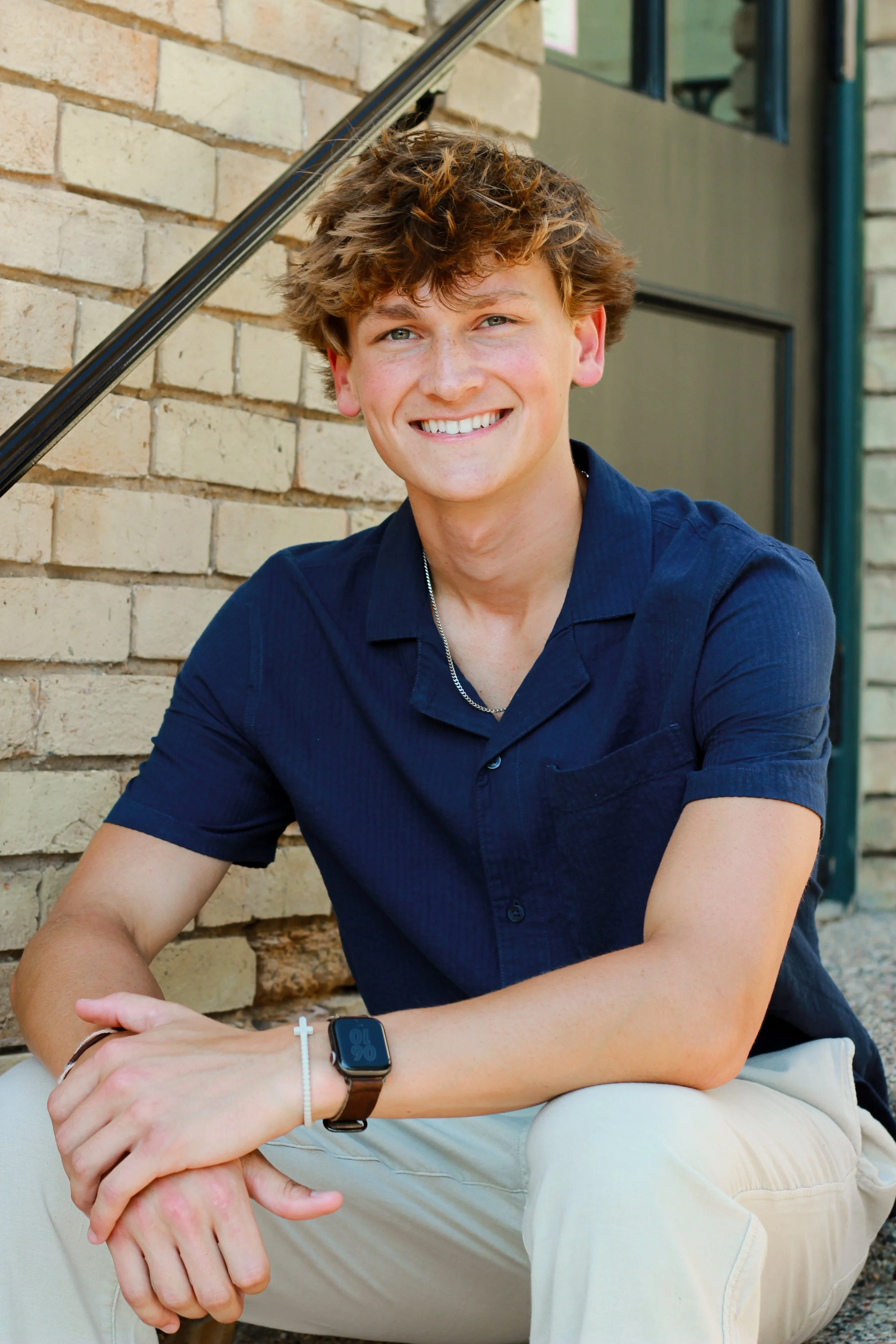 A young man with curly light brown hair, wearing a navy blue shirt and beige pants, sitting on the ground next to a brick wall, smiling at the camera.