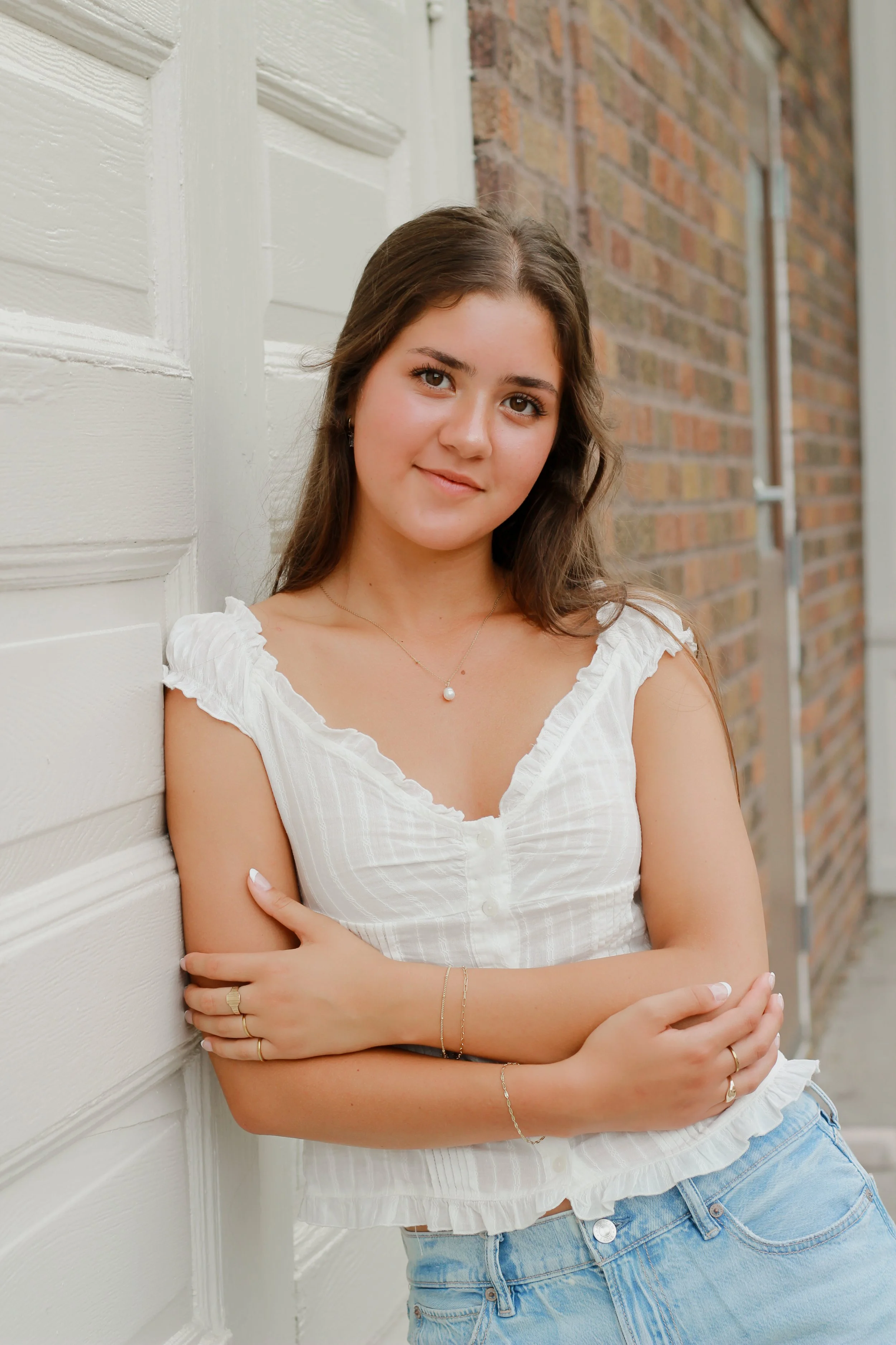 A young woman with long brown hair standing outdoors, leaning against a white wooden wall with brick background, wearing a white sleeveless top and denim shorts.