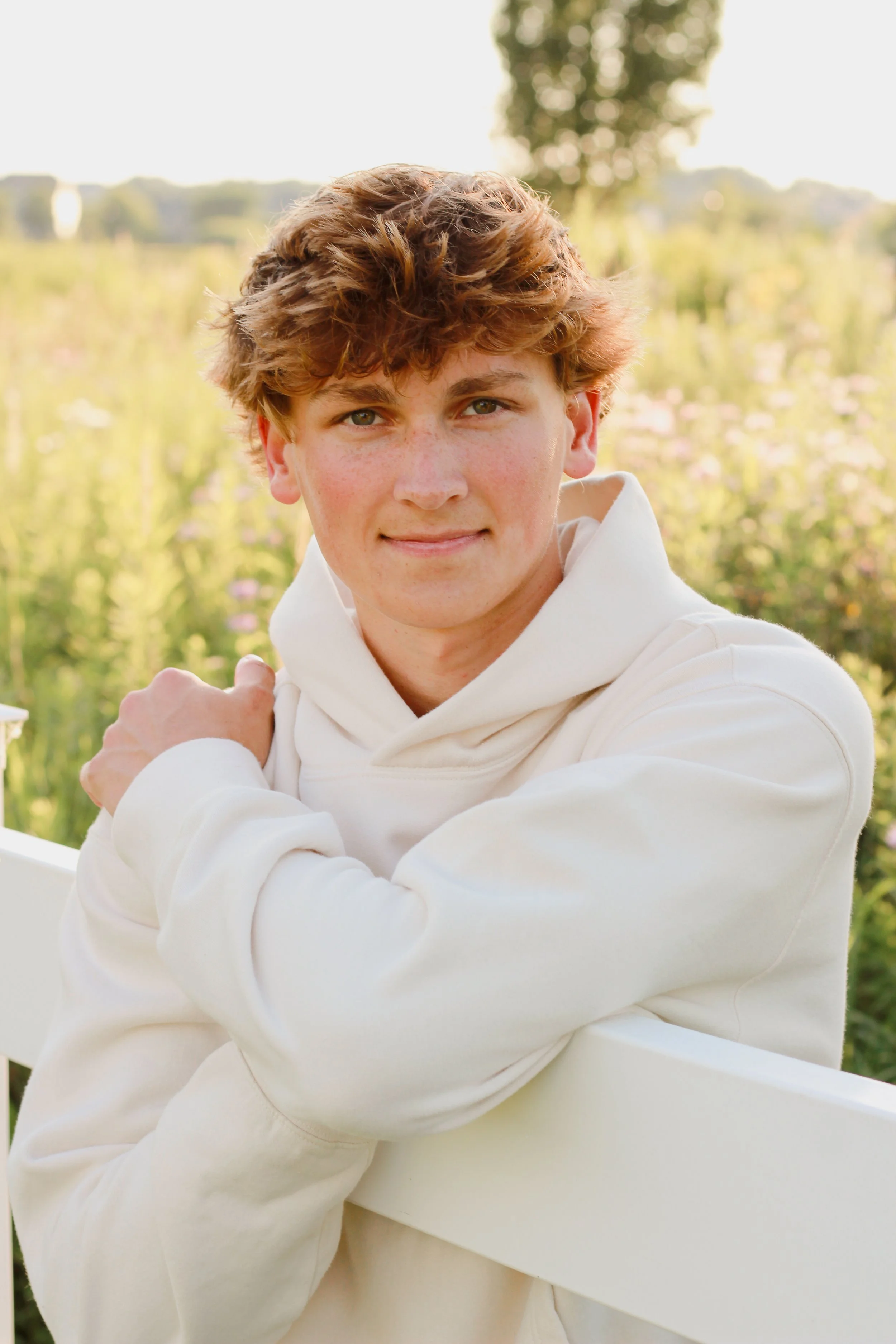 Young man with curly brown hair, wearing a white hoodie, standing outdoors in a field with greenery and flowers during daytime.