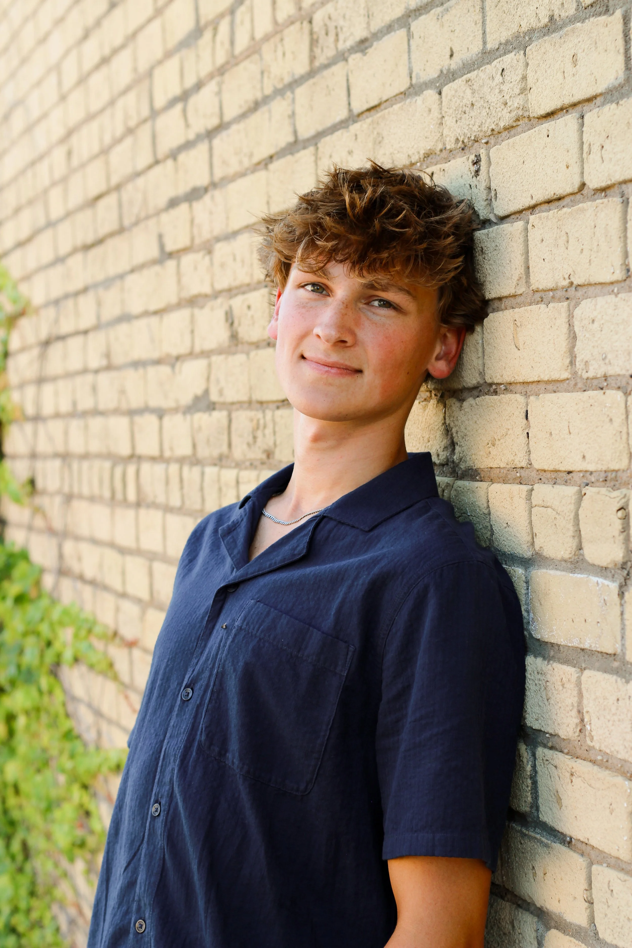 Young man with curly brown hair and light skin, wearing a navy blue shirt, leaning against a brick wall outdoors.