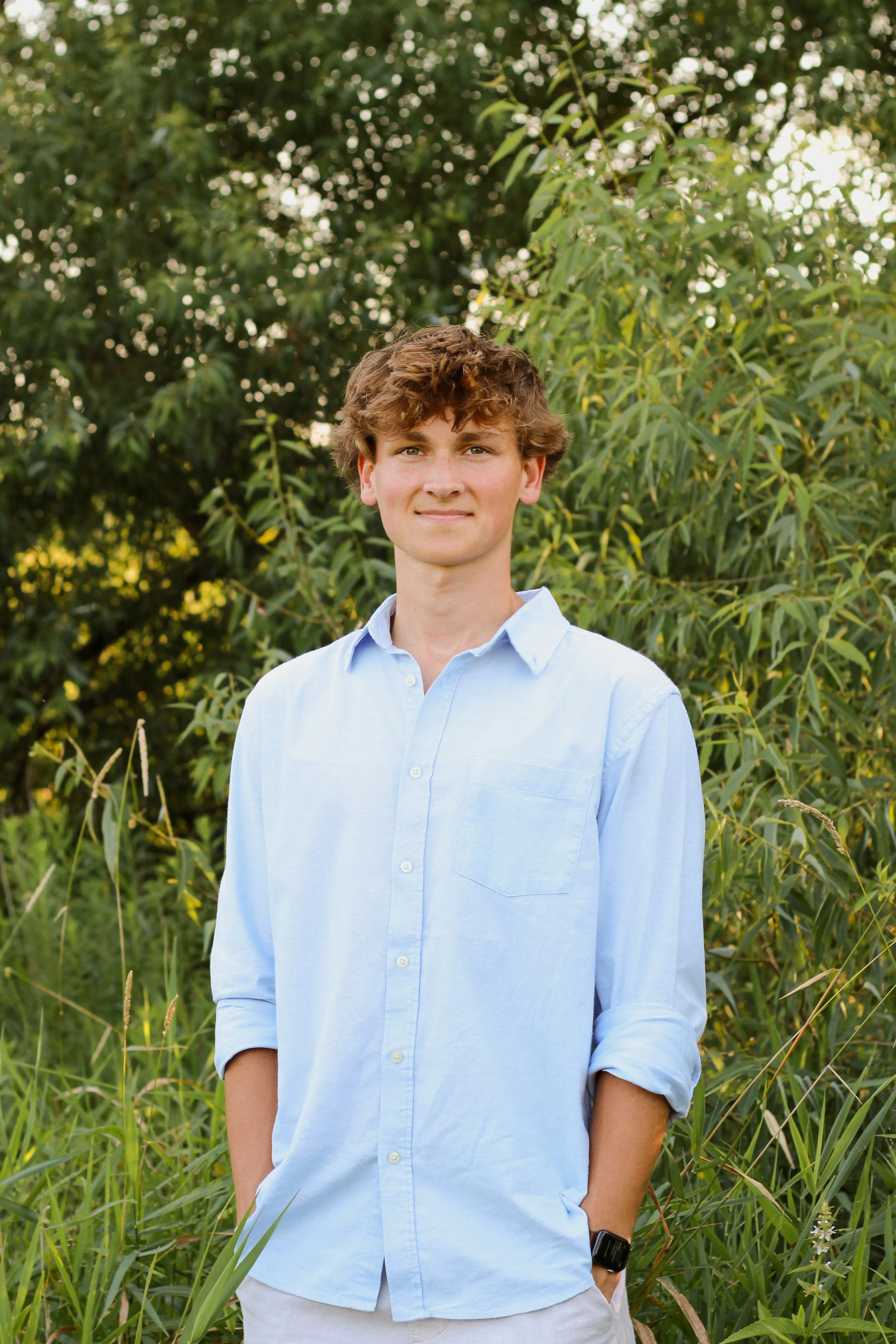 A young man with curly brown hair, wearing a light blue button-up shirt with rolled-up sleeves, standing outdoors in front of green trees and grass, with one hand in his pocket and a slight smile.