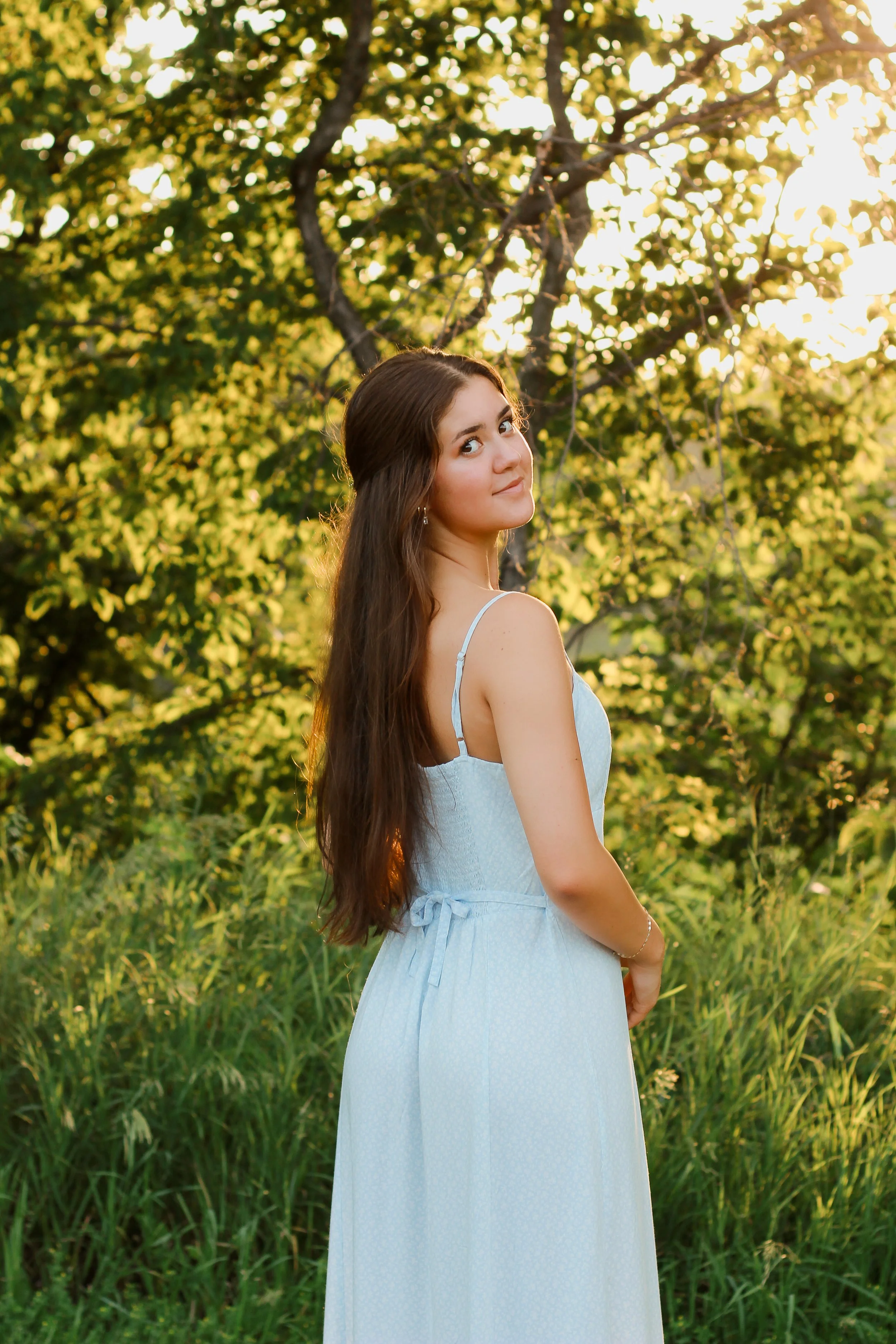 A young woman with long brown hair wearing a light blue dress standing outdoors in a grassy area with trees, during sunset.