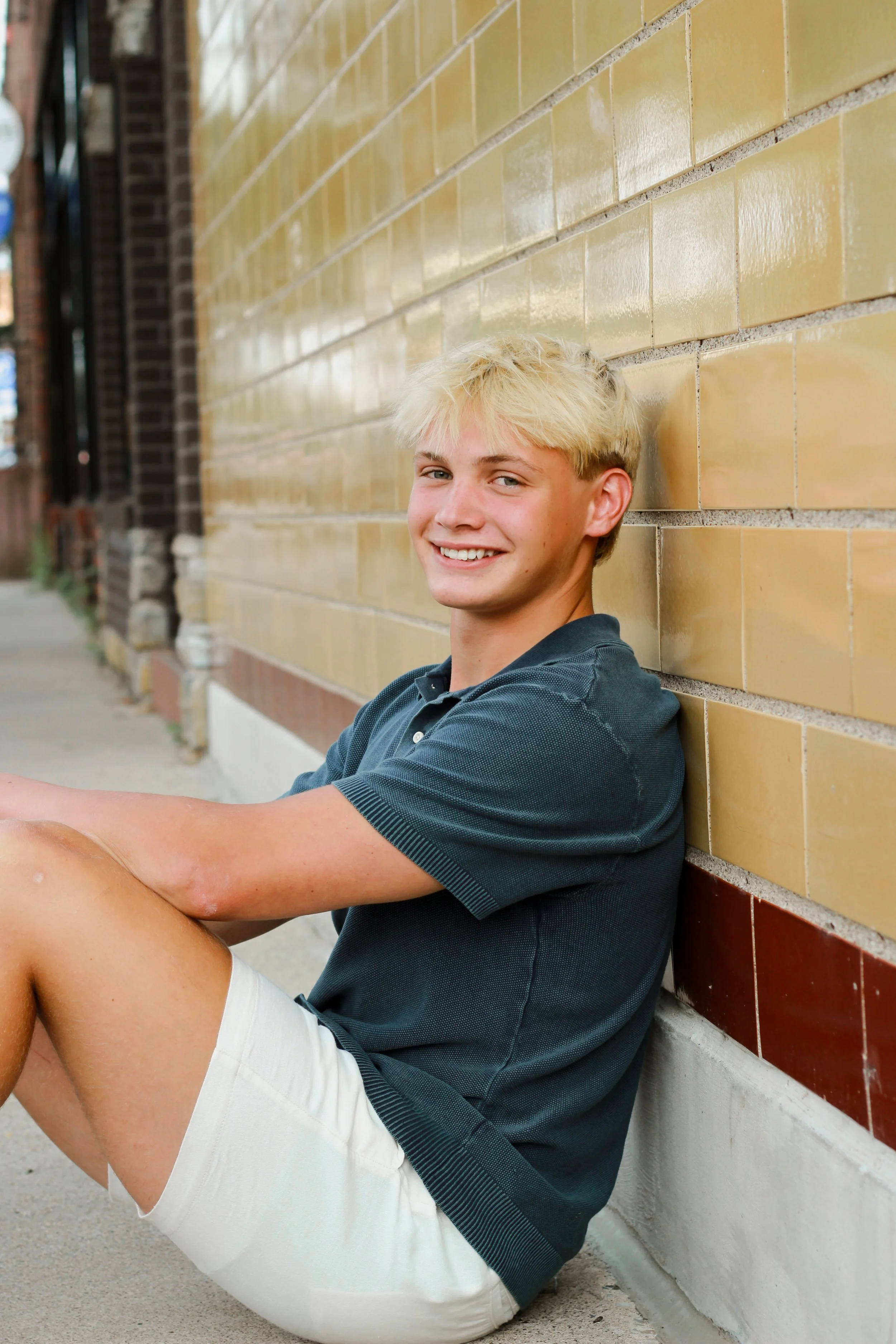 A teenage boy with blonde hair, wearing a dark polo shirt and beige shorts, sitting on the sidewalk next to a brick wall, smiling at the camera.