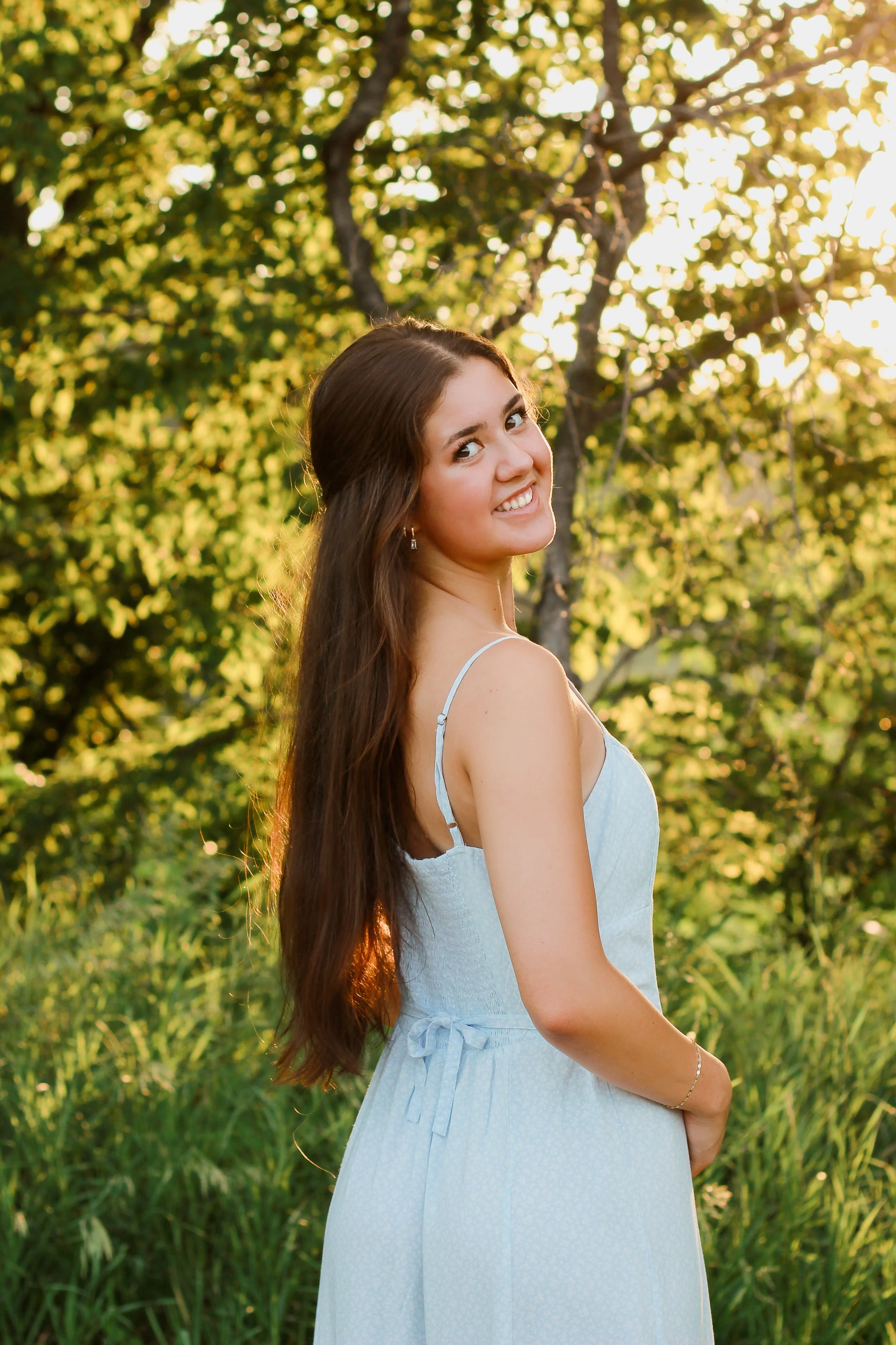 Young woman with long brown hair smiling in a light blue dress outdoors during sunset, with green trees and grass in the background.