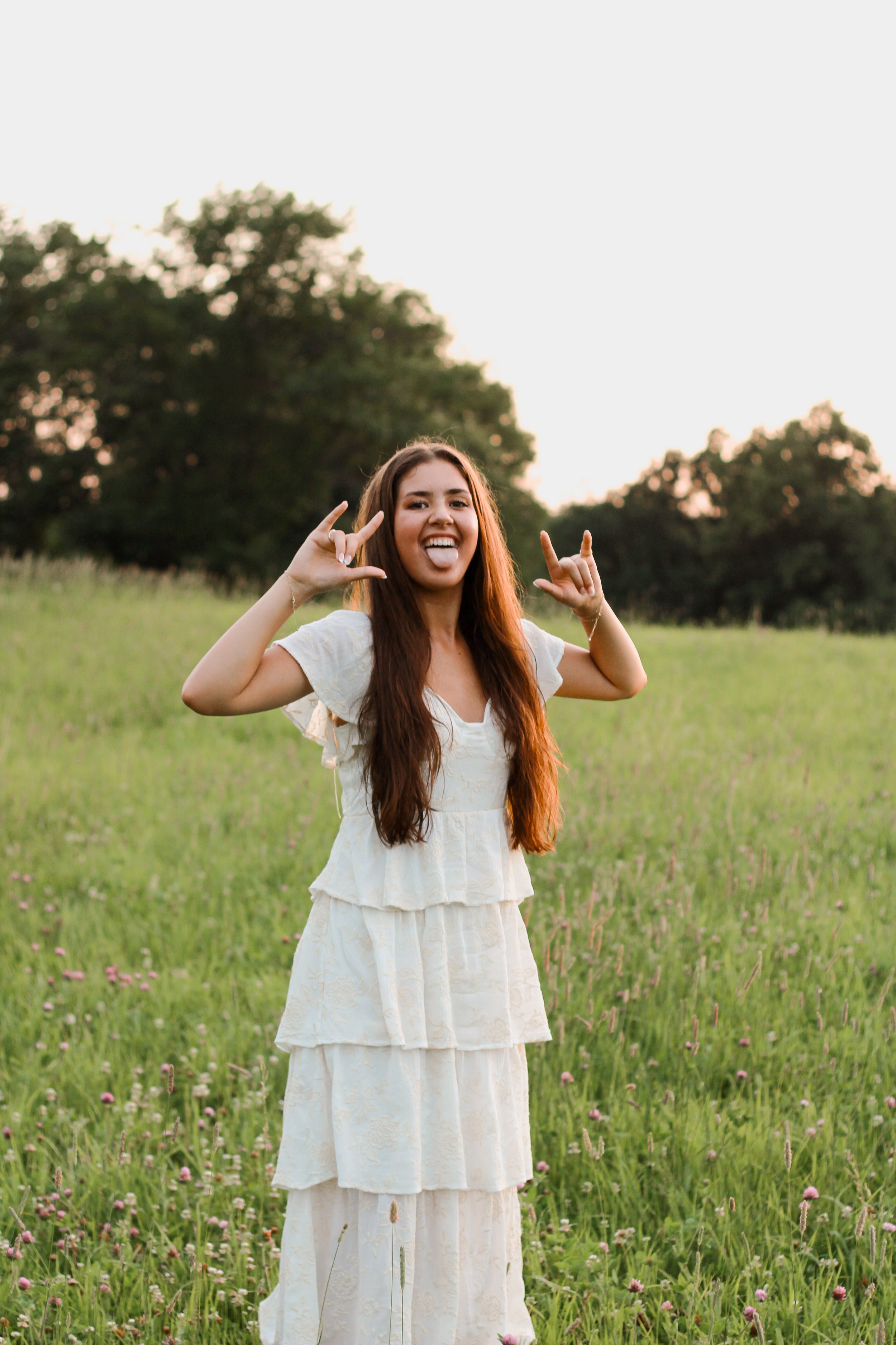 Young woman in a white dress making a rock and roll gesture in a green field during sunset with trees in the background.
