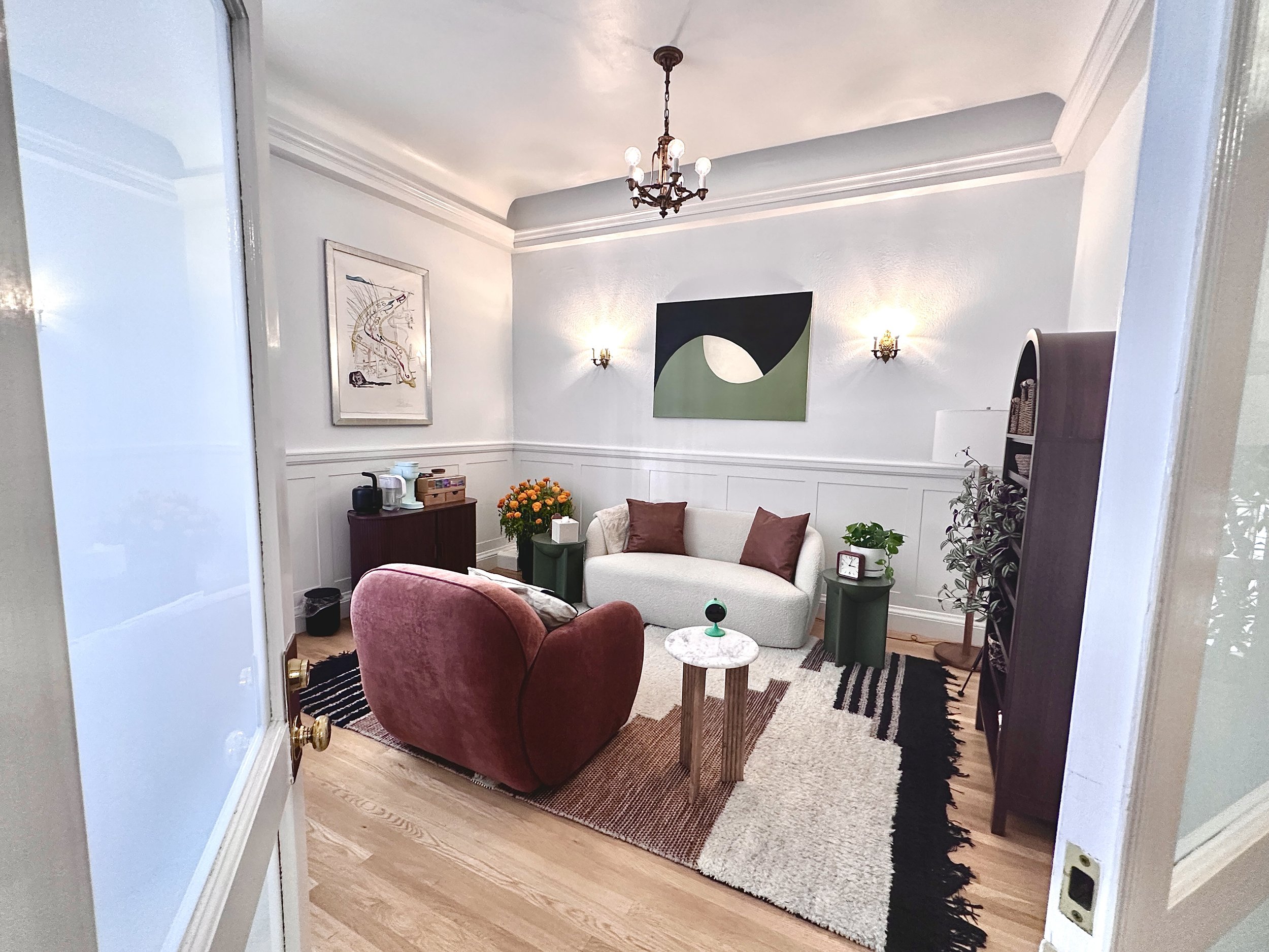 Living room with a chandelier, wall art, a white sofa with orange pillows, a maroon armchair, a side table with a small plant, a lamp, and decorative items, with a rug on wooden flooring.