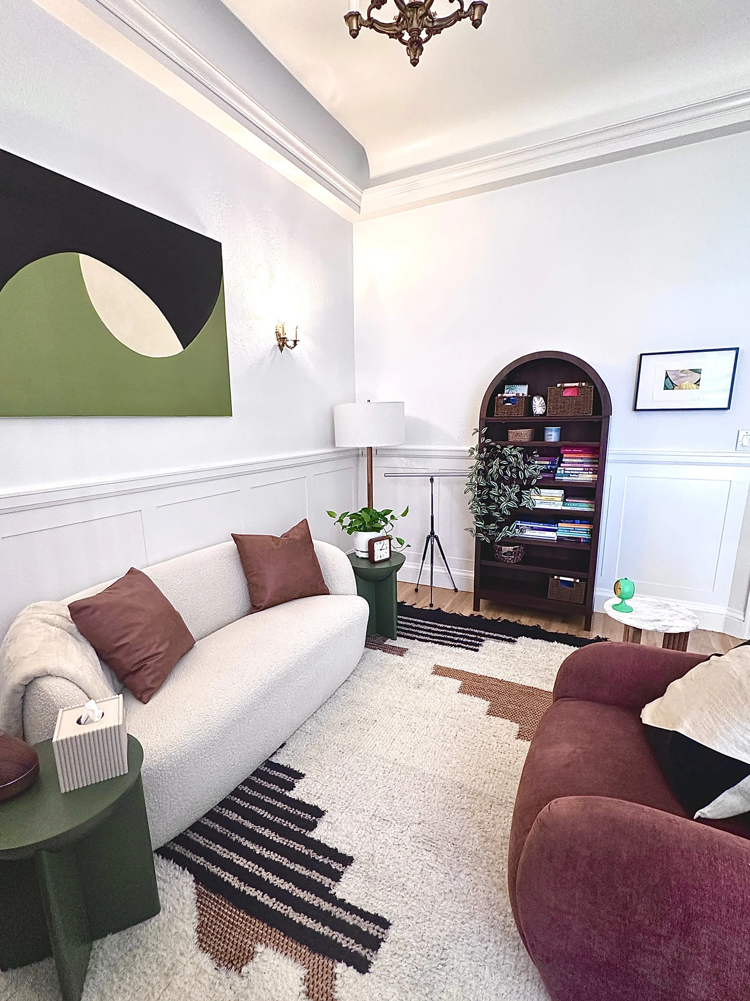 A cozy living room with a white sofa, brown pillows, a black and white striped rug, and a dark wooden bookcase filled with books and decorative items. There is a white lamp, a small potted plant, and a marble-topped side table with a plant and a green lamp. The walls are painted white with artwork hanging, and a chandelier is visible on the ceiling.