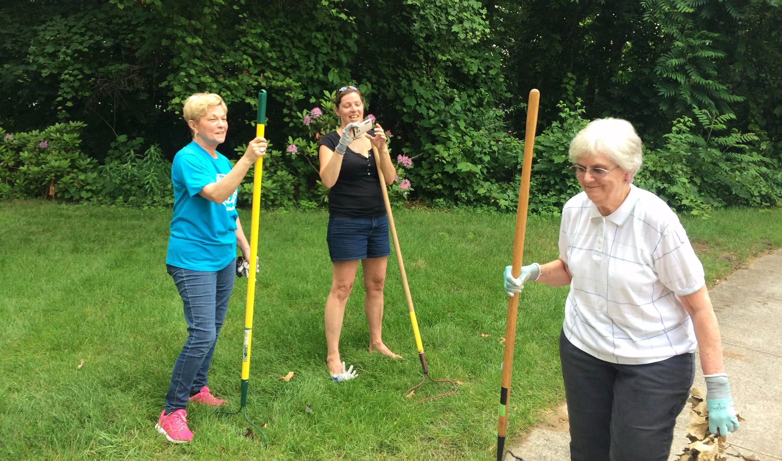 Three women are working outdoors on a gardening project, with one woman holding a gardening tool and another taking a photo, while the third woman is walking on a paved path.