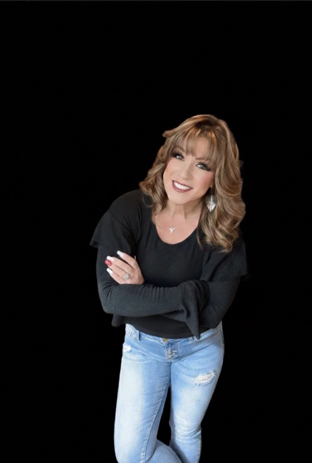 A woman with light brown, shoulder-length hair and styled bangs, wearing a black long-sleeve shirt and ripped blue jeans, smiling and posing with her arms crossed against a black background.