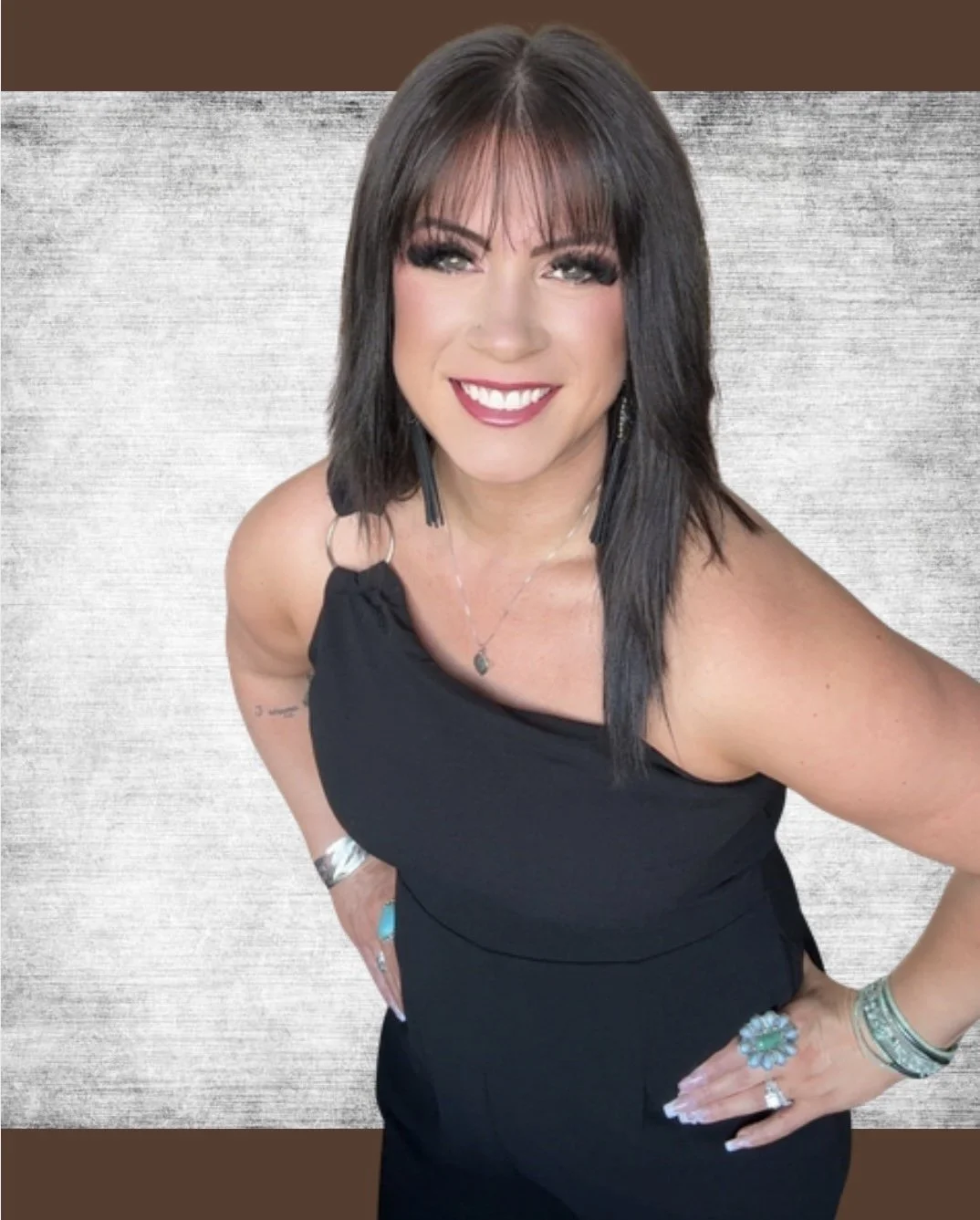 A smiling woman with shoulder-length dark brown hair, wearing a black sleeveless dress, jewelry including rings, bracelets, earrings, and a necklace, standing against a textured background.