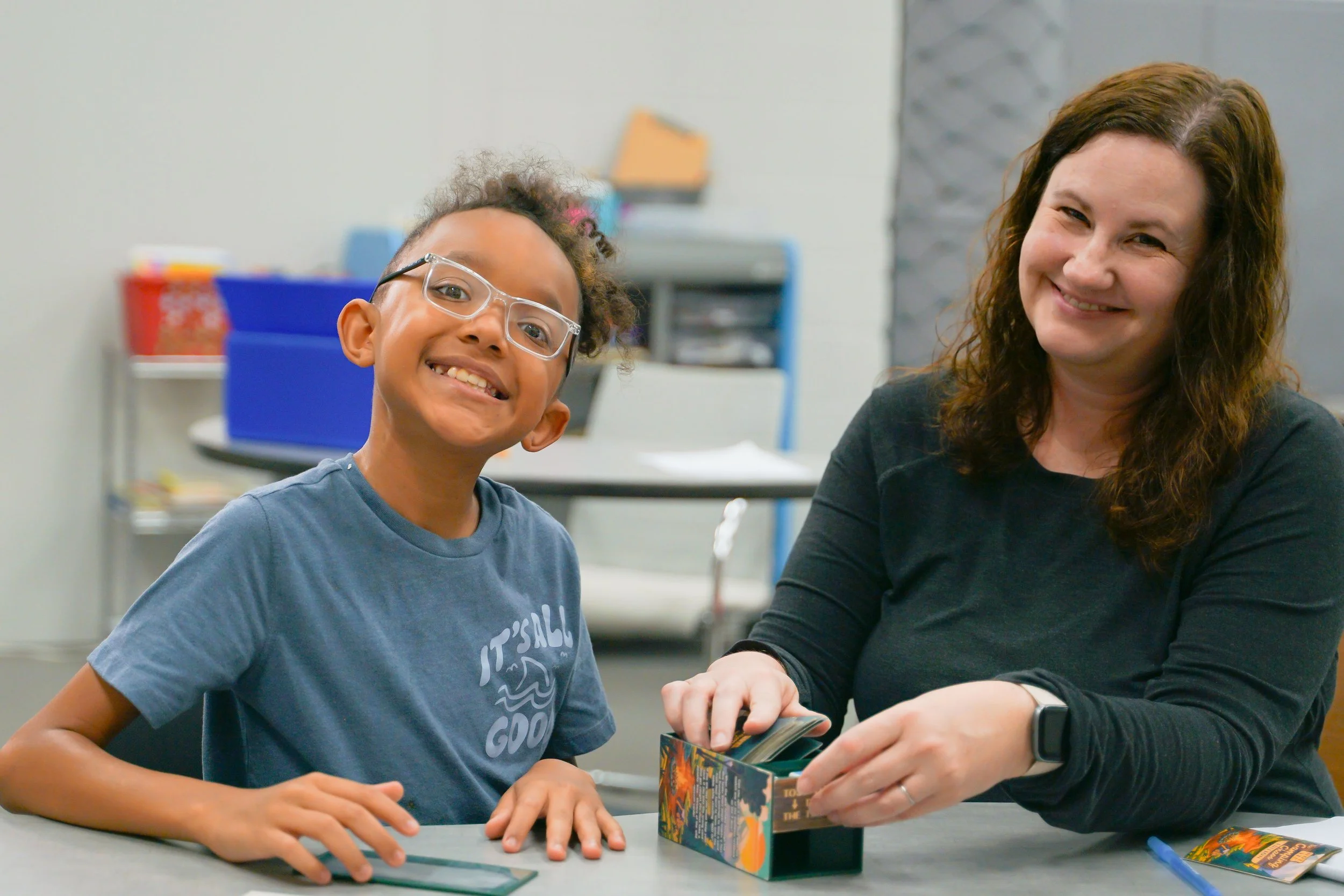 A smiling boy with glasses and a woman sitting at a table, playing a card game together in a classroom or recreational room.