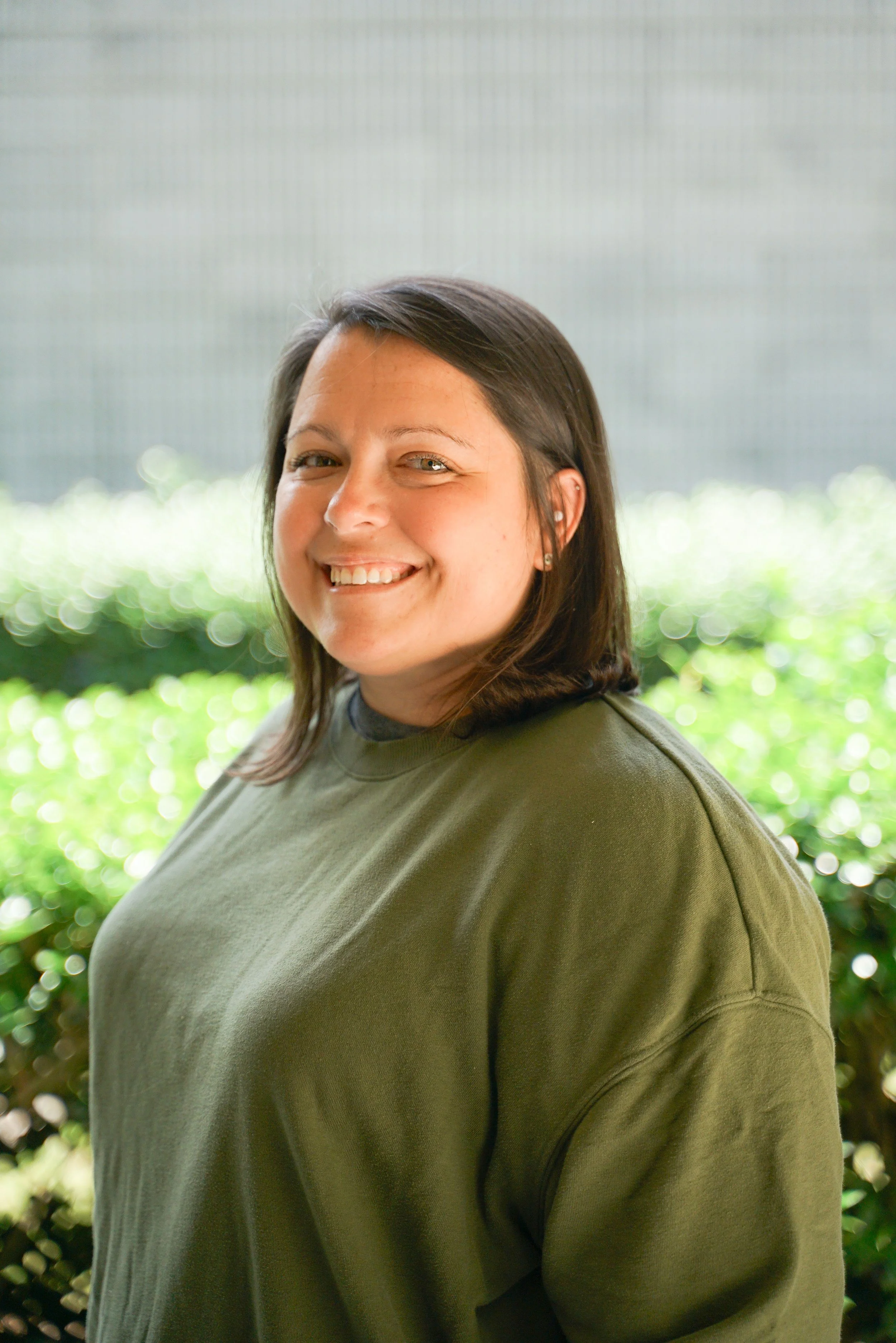 A smiling woman with shoulder-length brown hair, wearing a green sweatshirt, standing outdoors with green bushes and a gray background.