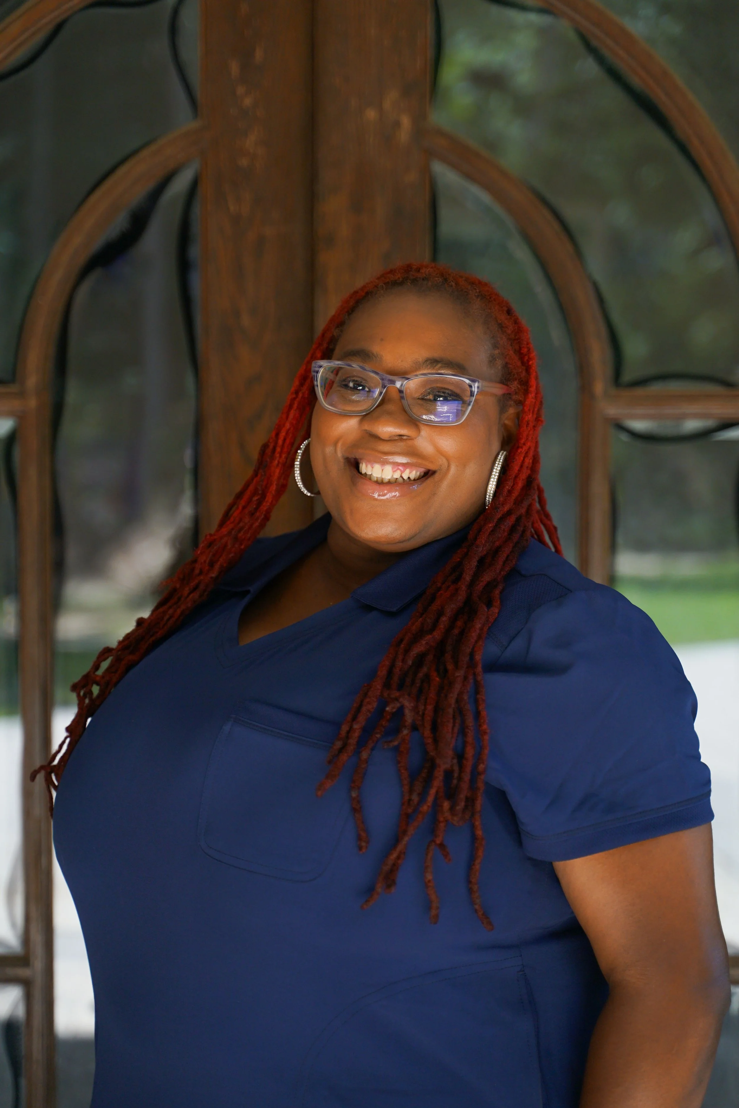 A smiling woman with red dreadlocks, wearing glasses, hoop earrings, and a navy blue shirt, standing in front of a wooden window frame.