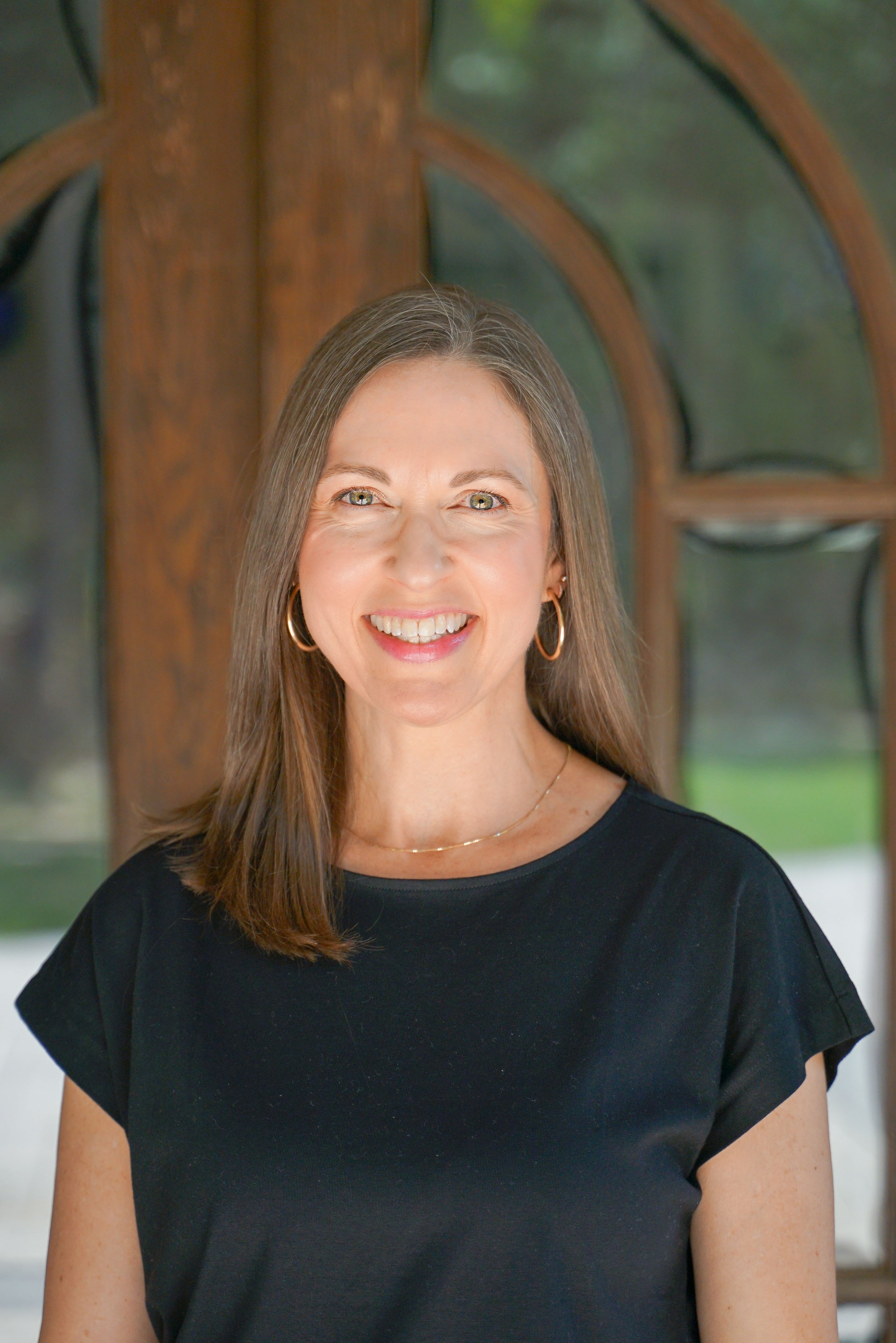 A woman with long brown hair wearing a black top, gold hoop earrings, and a delicate gold necklace. She is smiling and standing in front of a wooden archway with glass panes, with a blurred outdoor background.