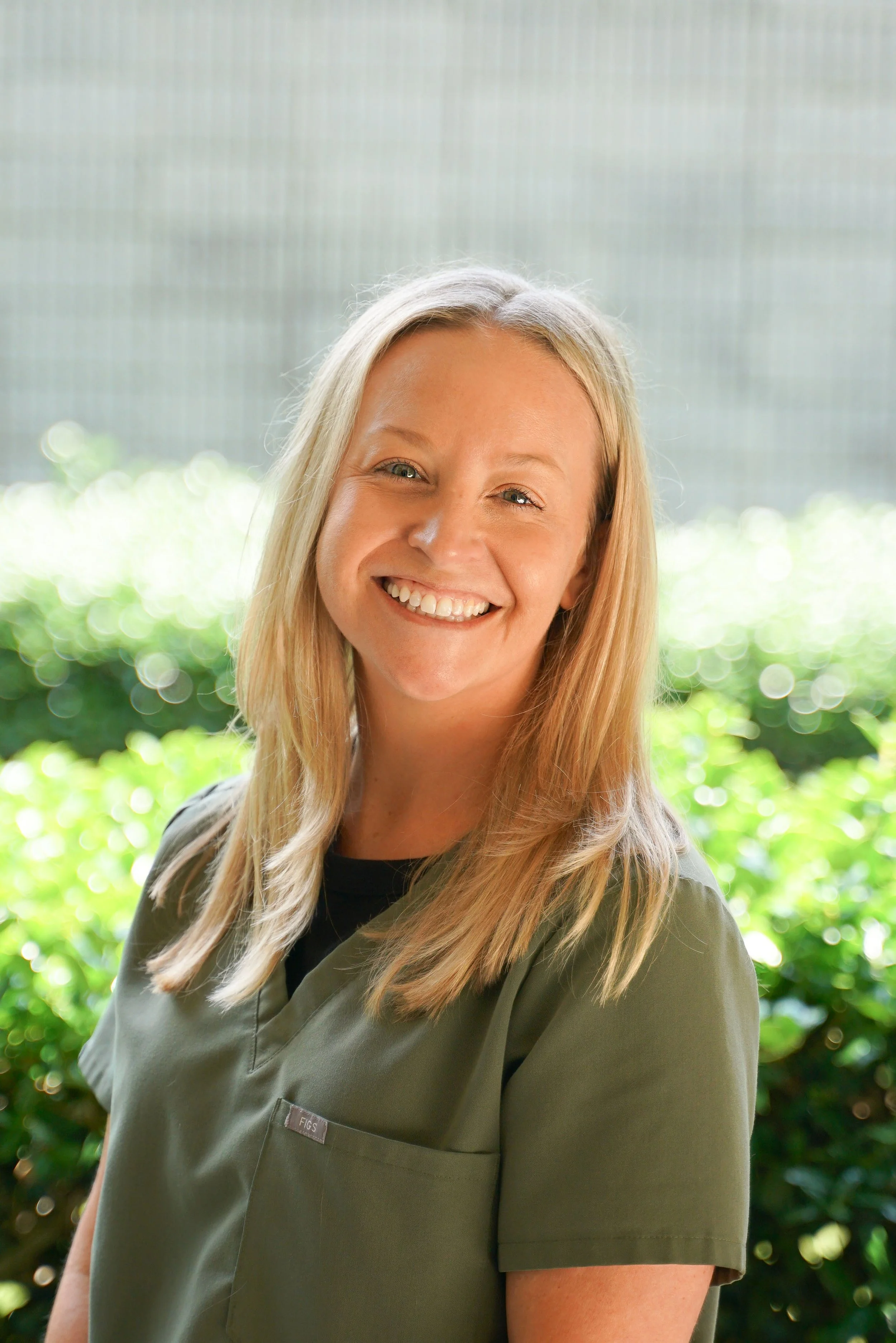 A smiling woman with blonde hair wearing a green medical uniform outdoors with greenery in the background.
