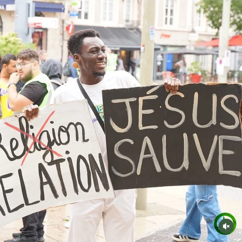 Un homme tient deux pancartes lors d'une manifestation. La première indique 'Religions' avec un croisé sur le mot, et la seconde dit 'Jésus sauve'.