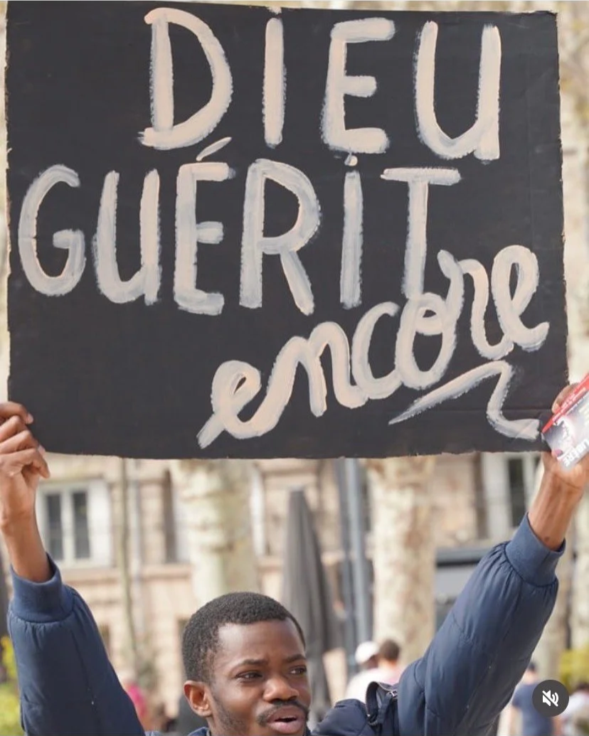 Un homme tient une pancarte noire avec l'inscription en blanc : "Dieu guérit, envoie" dans une manifestation en extérieur.