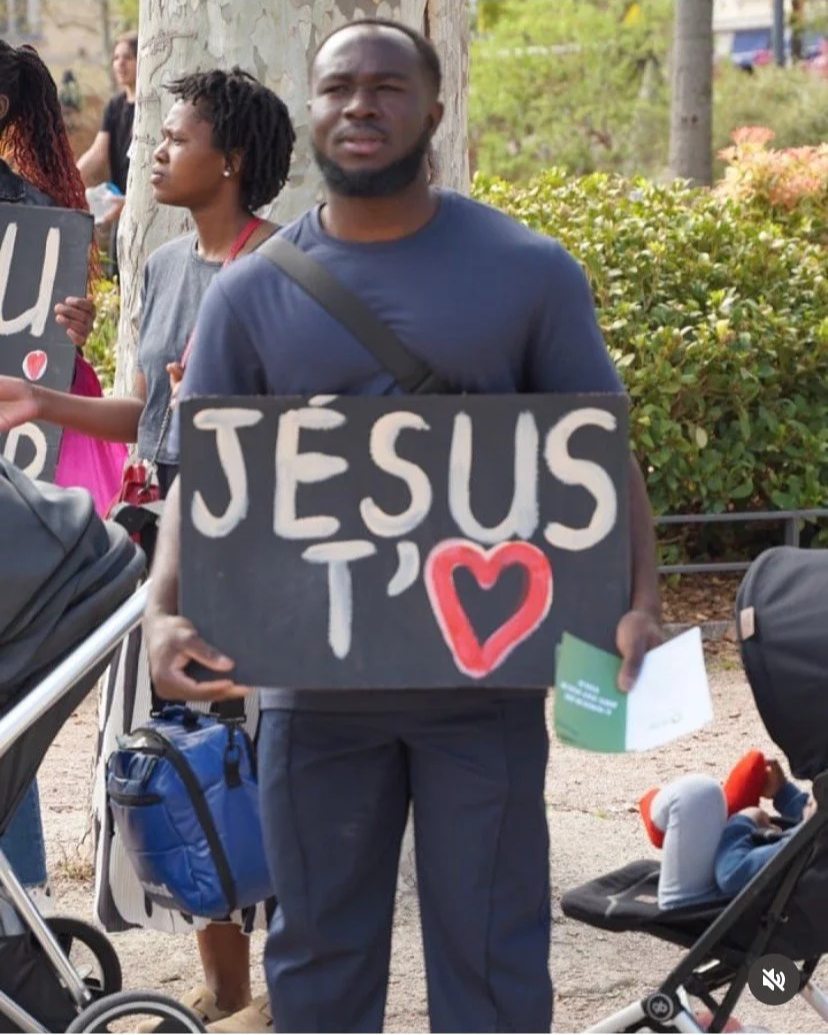 Homme tenant une pancarte noire avec l'inscription "JÉSUS T ❤" en lettres blanches et rouges, dans un parc.
